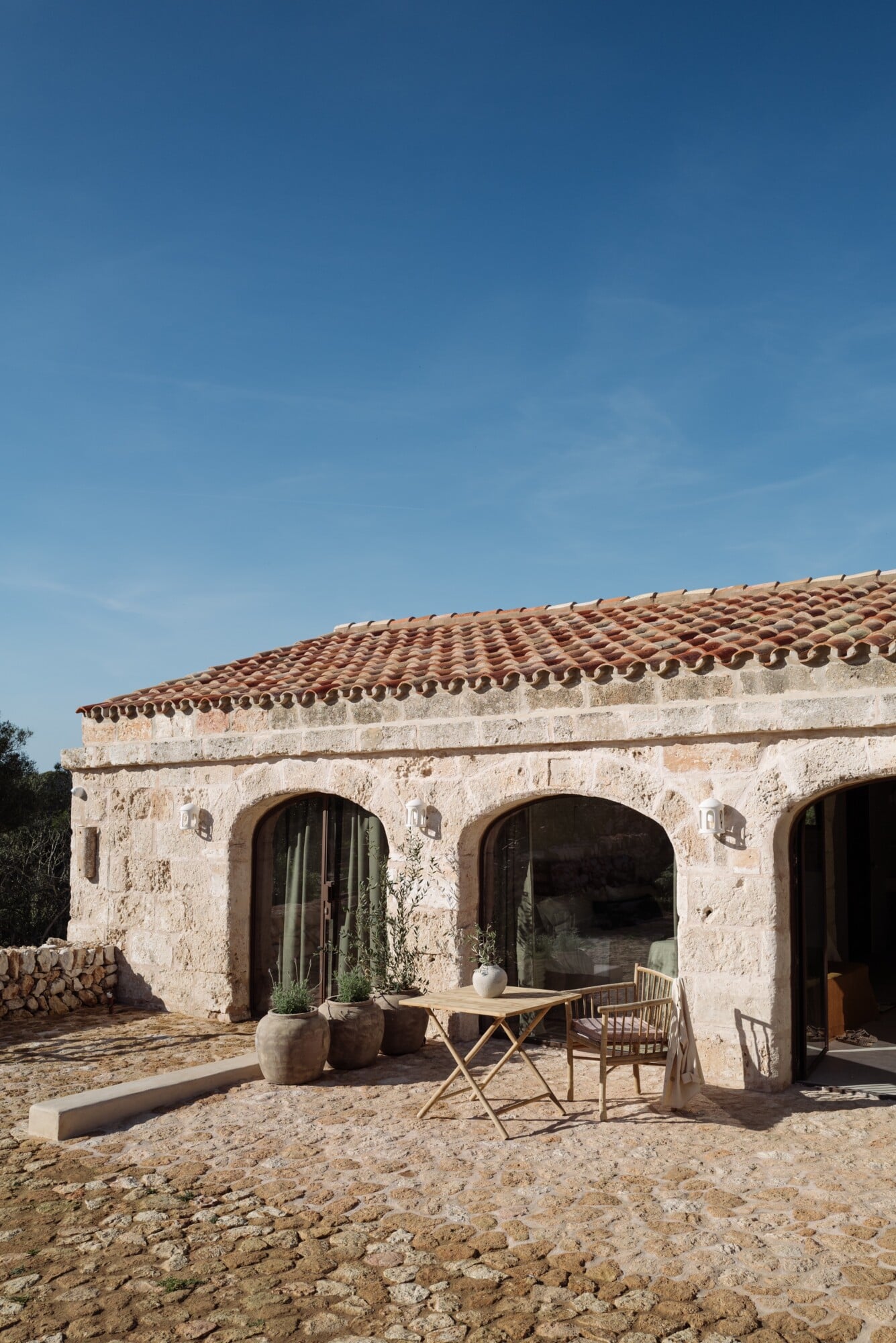 Santa Ana, Balearic Islands — Terracotta-roofed stone villa with arched openings, minimalist outdoor furniture, and sun-bleached courtyard under clear sky.