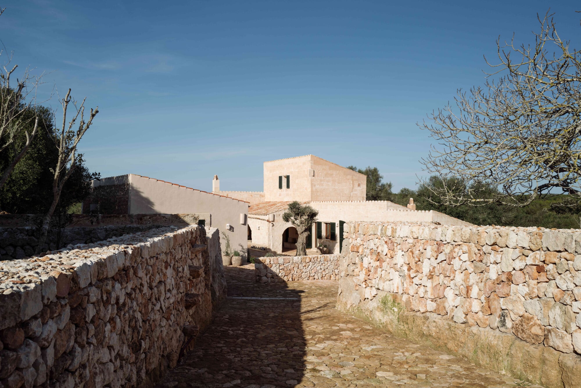Santa Ana, Balearic Islands — Whitewashed Mediterranean villa with stone courtyard, arched entryway, and terraced limestone walls under clear sky.