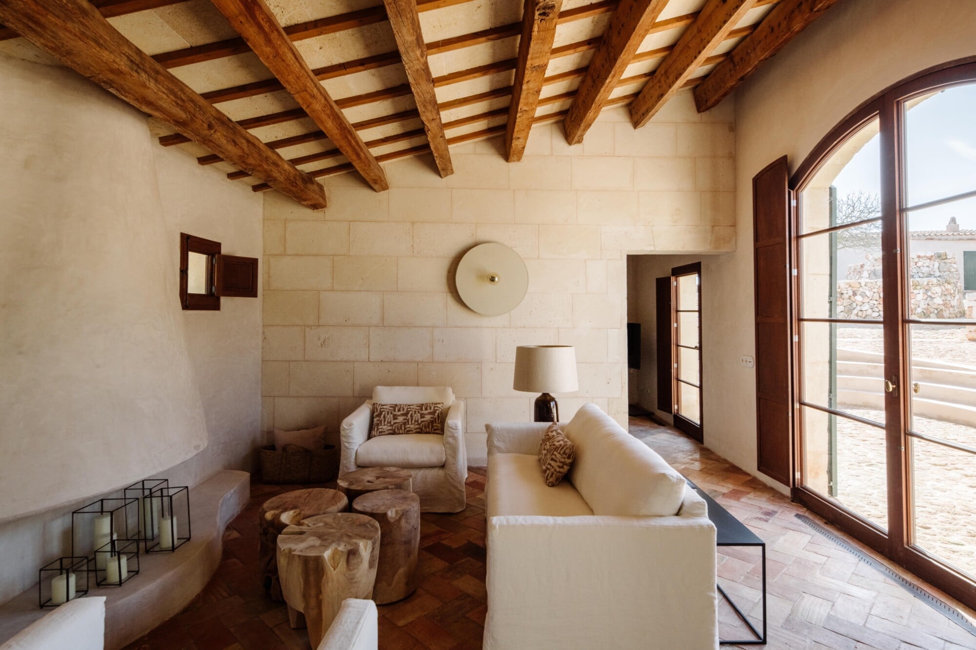 Santa Ana, Balearic Islands — Minimalist living room with exposed wooden beam ceiling, cream stone walls, linen seating, wooden stools, and large arched doors opening to terrace.