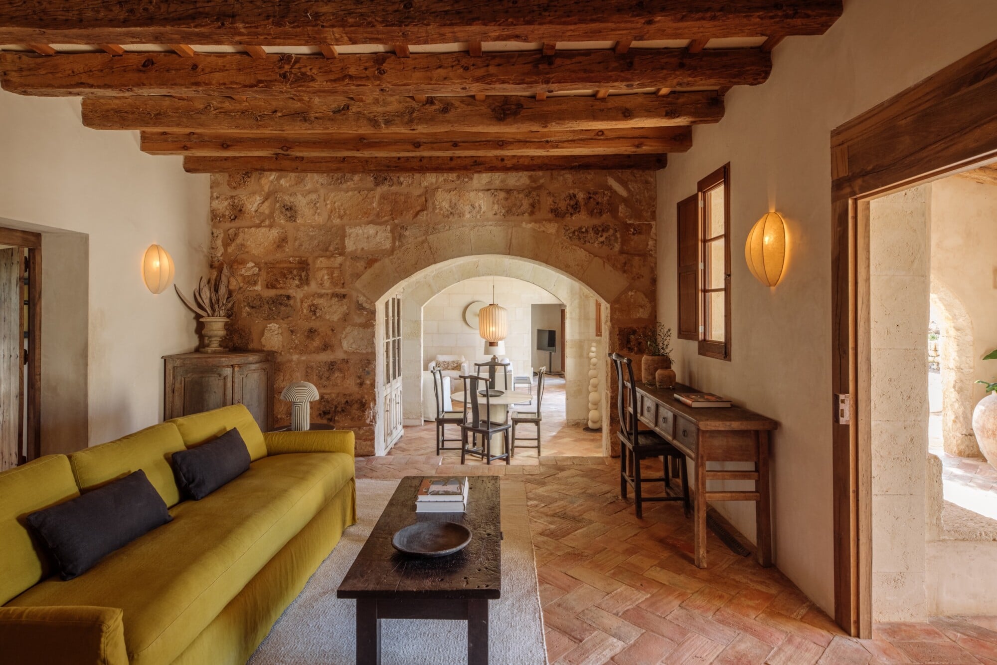 Santa Ana, Balearic Islands — Arched stone passageway connecting ochre-walled living room with mustard sofa to dining area, exposed wood beams, terracotta flooring, pendant lights.