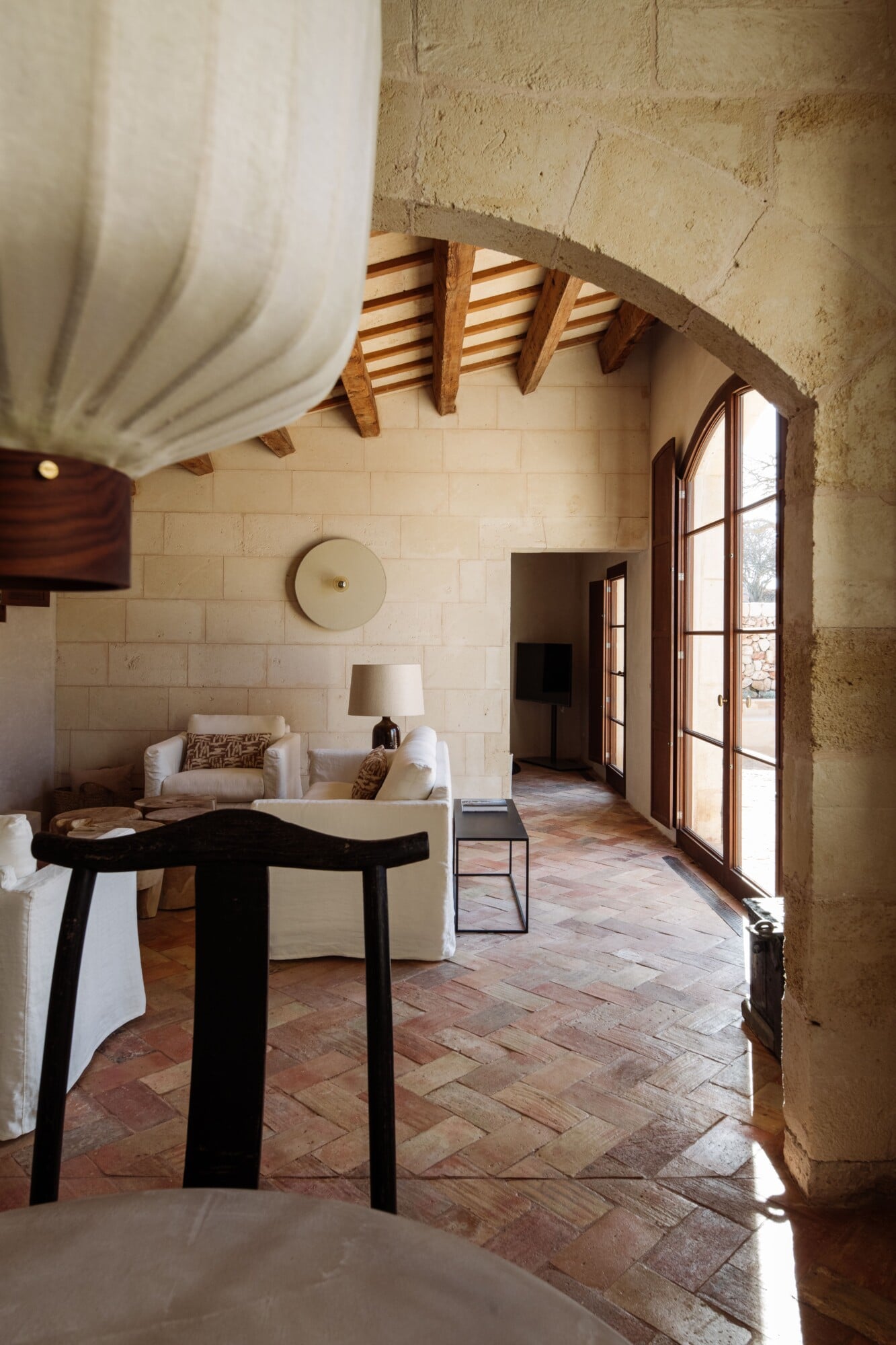Santa Ana, Balearic Islands — Stone archway, wooden beam ceiling, terracotta herringbone floors, cream furnishings, natural light from tall windows in minimalist Mediterranean interior.