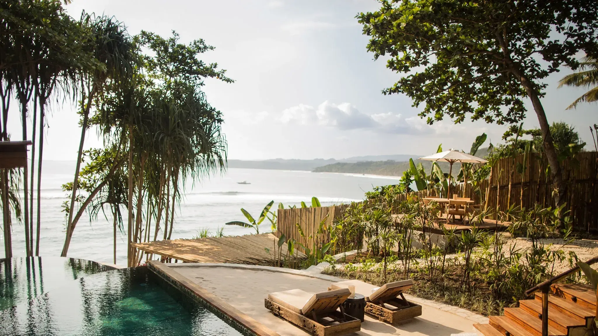 NIHI Sumba, Asia — Infinity pool overlooking coastal bay with teak loungers, wooden deck, banana plants, and palm trees framing turquoise waters under overcast sky.