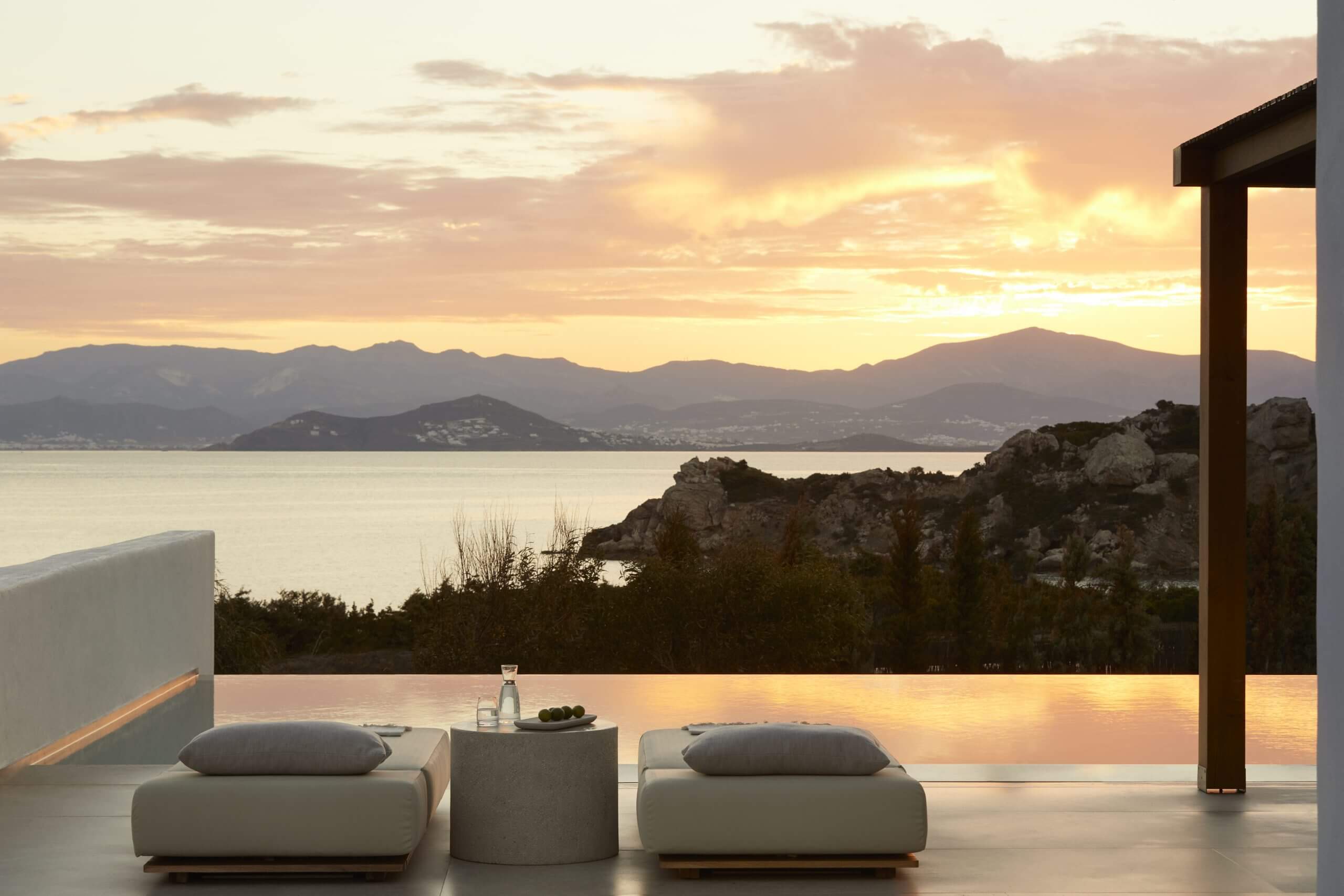 Pārocks, Europe — Minimalist poolside lounge at dusk with limestone seating, panoramic water views, distant mountains, and golden hour lighting across infinity pool.
