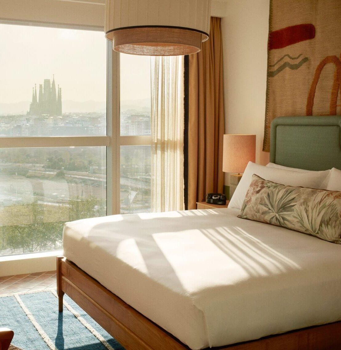 The Hoxton Poblenou, Barcelona, Spain — Sunlit bedroom with sage headboard, linen bedding, pendant lamp, and Sagrada Familia views through floor-to-ceiling windows.