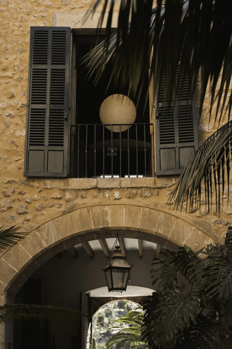 Hotel Corazón, Balearic Islands — Ochre stone facade with dark wooden shutters, black wrought-iron balcony, arched entryway, and palm shadow pattern in Mediterranean sunlight.