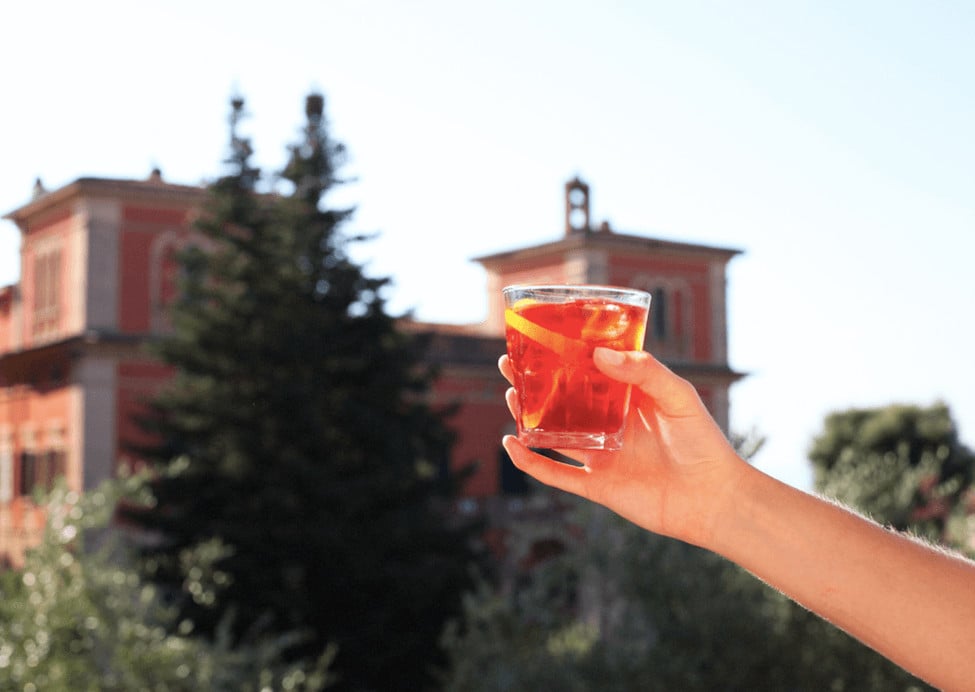 Aperitivo Bar — Hand holding coral-red aperitivo cocktail with terracotta villa and cypress trees, European countryside aesthetic, warm golden hour light.