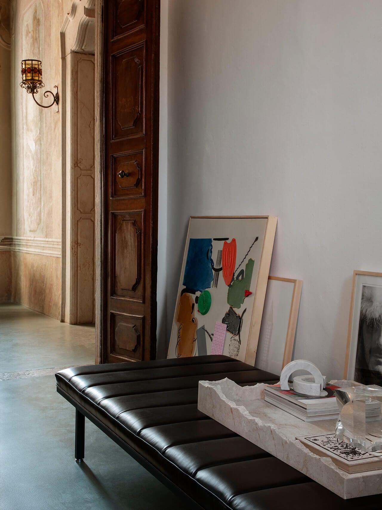 Vipp Palazzo Monti, Italy — Black leather bench in minimalist gallery space with travertine table, artworks, ornate wooden doors, and classical architecture.
