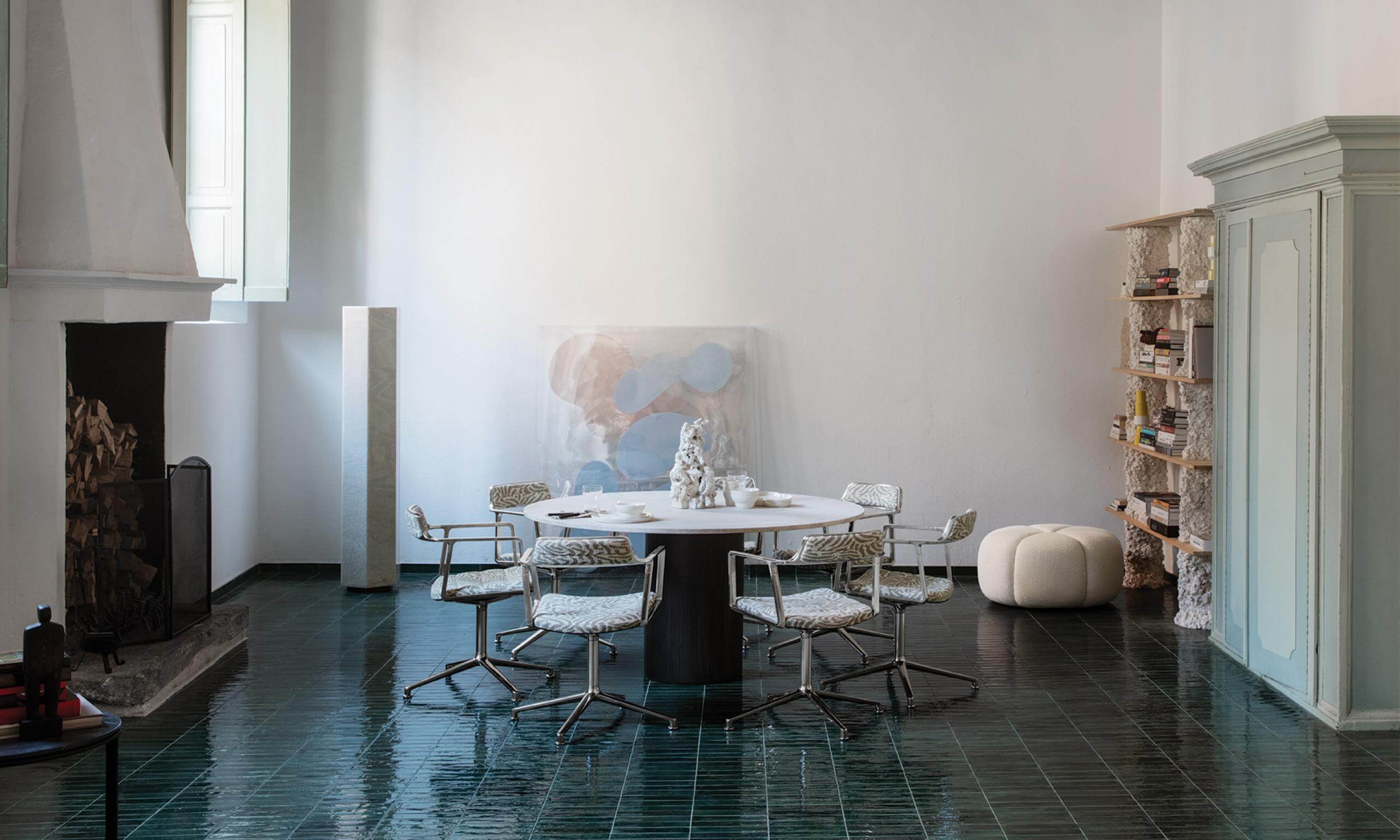 Vipp Palazzo Monti, Italy — Minimalist dining space with round black table, patterned chairs, pale stone pouf, and gallery artwork against white walls.