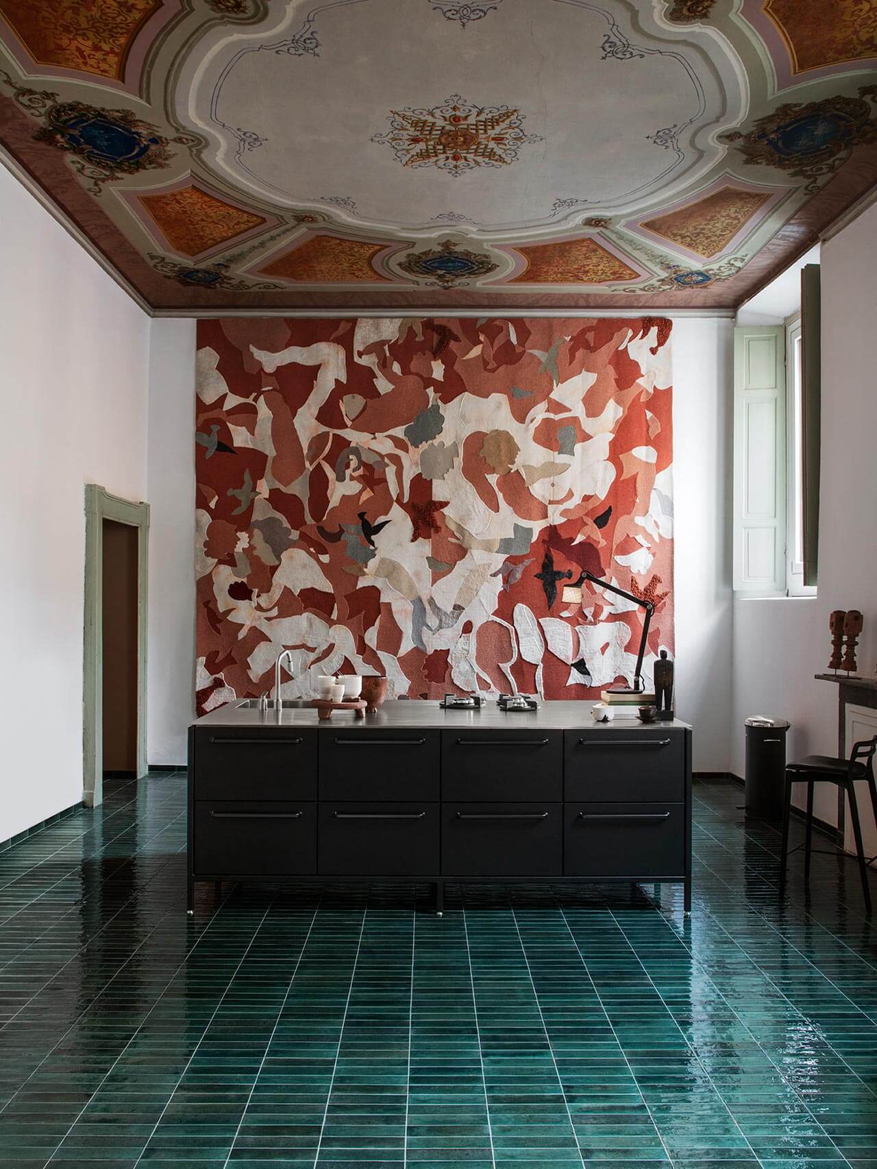 Vipp Palazzo Monti, Italy — Minimalist black cabinetry beneath abstract red-and-white tapestry, ornate stucco ceiling, green tile flooring, gallery aesthetic.