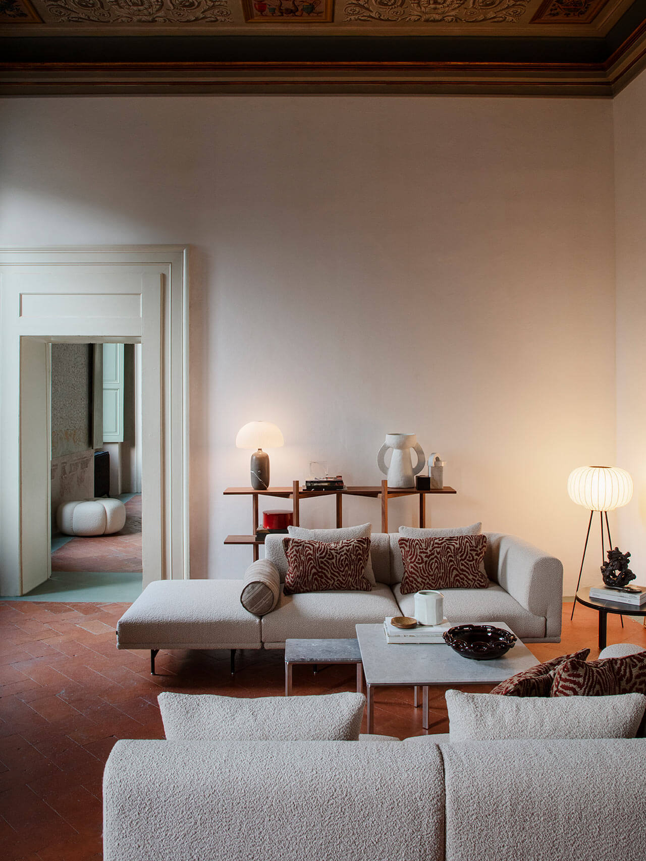 Vipp Palazzo Monti, Italy — Minimalist living room with beige sectional sofa, wooden shelving, pendant lamps, terracotta floors, and ornate ceiling detail.