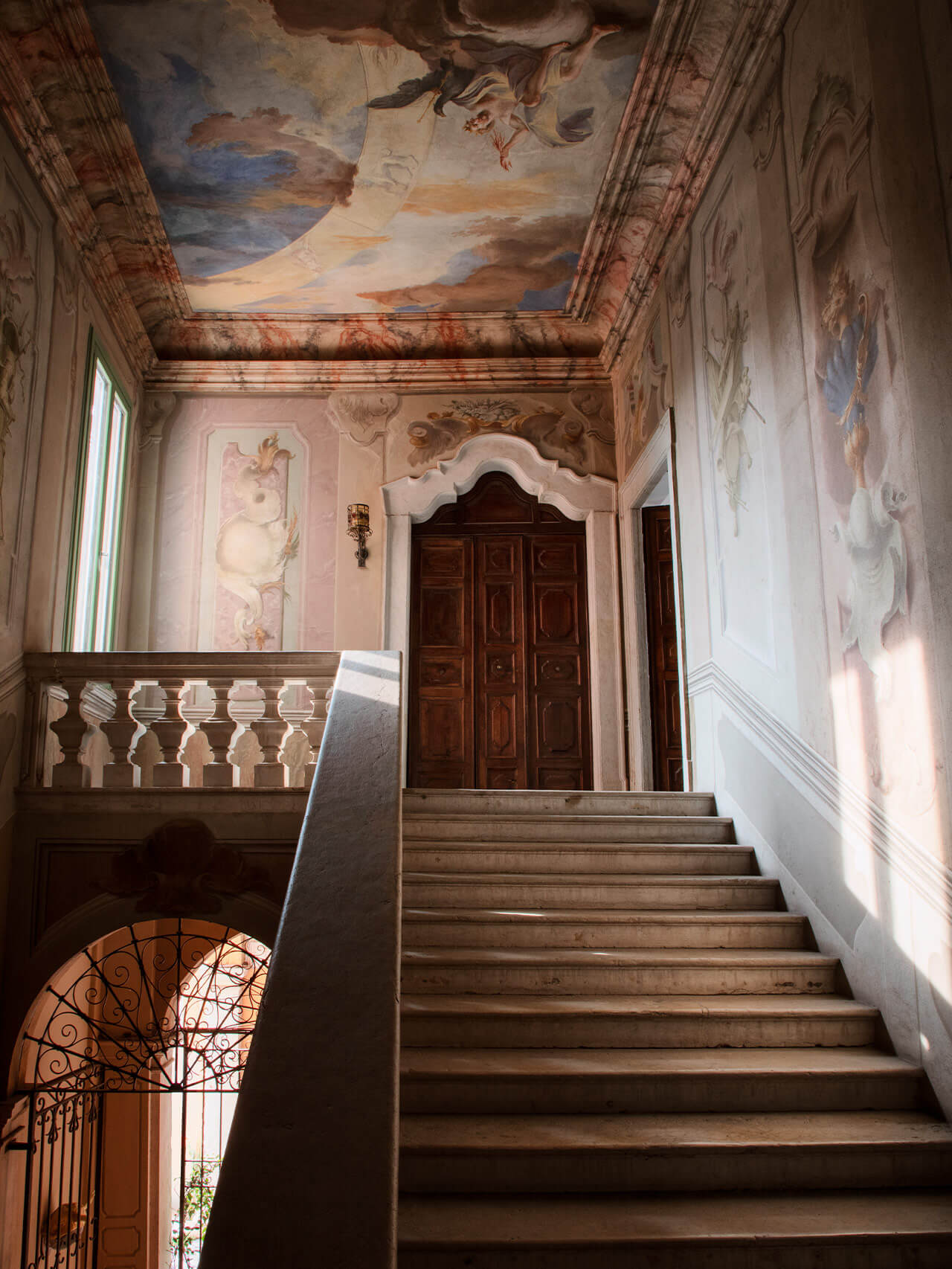 Vipp Palazzo Monti, Italy — Neoclassical staircase with frescoed ceiling, ornamental balustrade, wooden doors, and warm natural light illuminating terracotta walls.