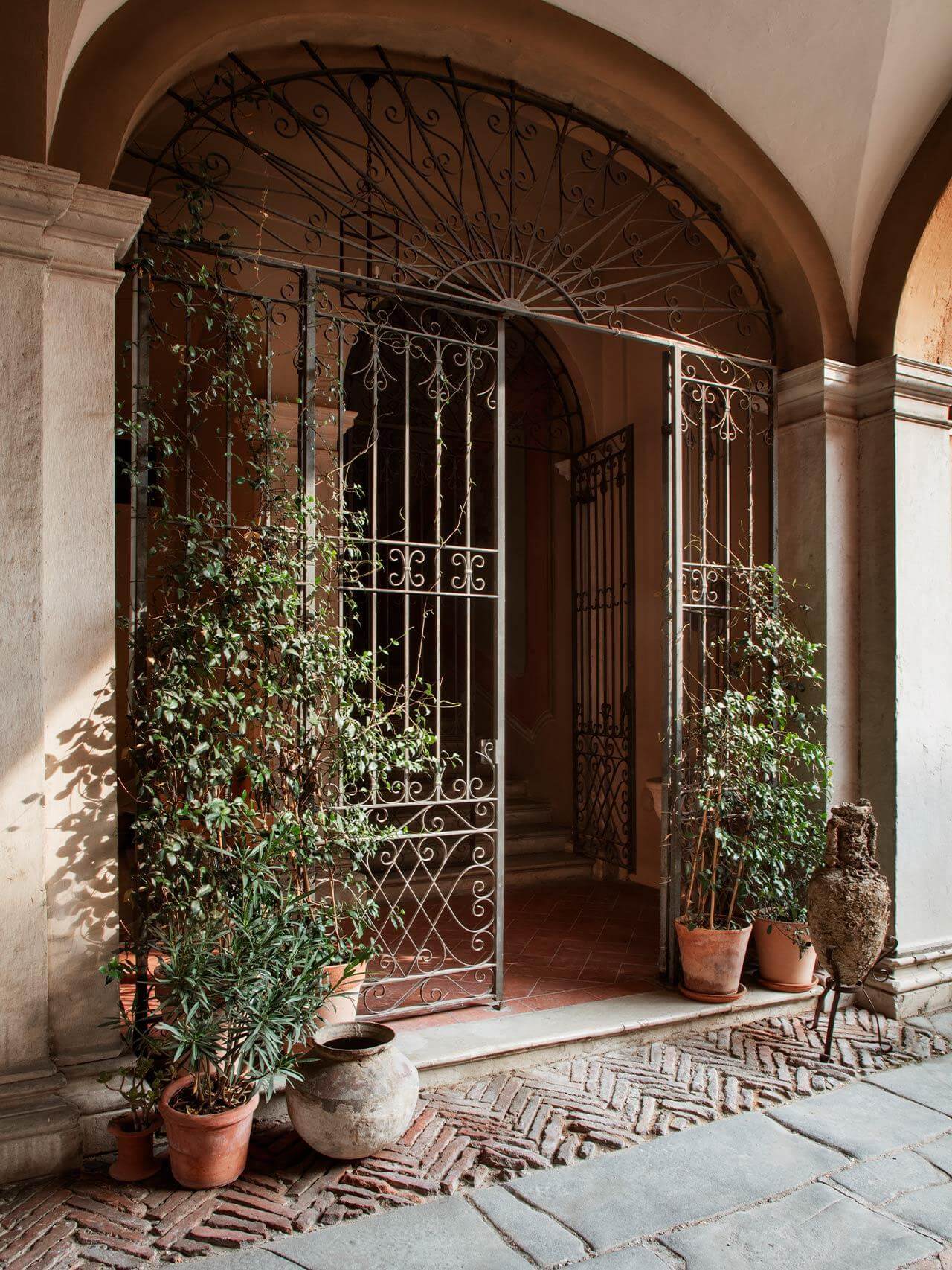 Vipp Palazzo Monti, Italy — Arched stone corridor with ornate wrought-iron doors, climbing ivy, terracotta pots, and warm natural light casting shadows on herringbone brick floor.