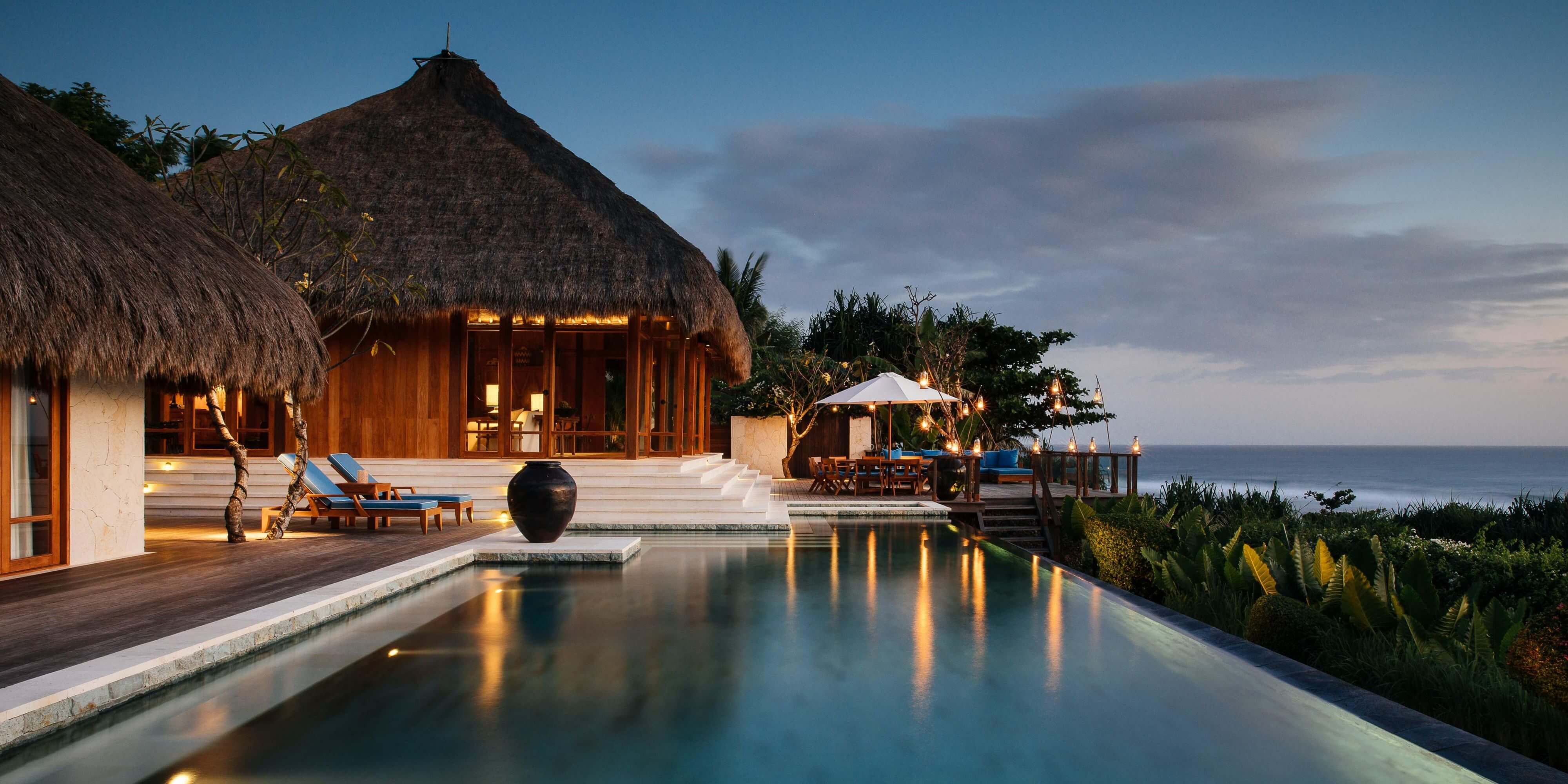 NIHI Sumba, Indonesia — Clifftop infinity pool at dusk with thatched-roof pavilion, wooden loungers, and ocean horizon beyond.