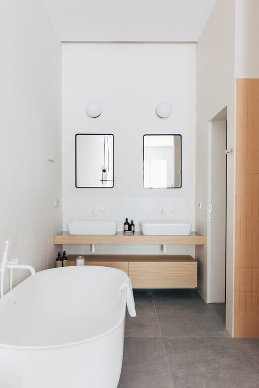 Plácido y Grata, Europe — Minimalist bathroom with dual vessel sinks, black-framed mirrors, natural wood vanity, white soaking tub, and concrete flooring.