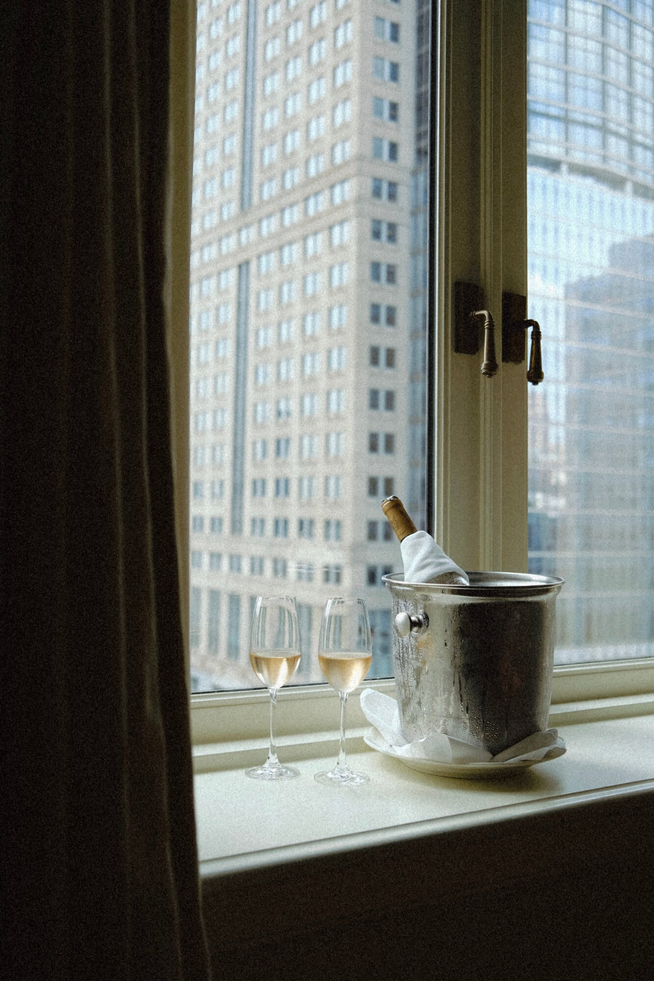 The Greenwich Hotel, New York, USA — Champagne flutes and ice bucket on windowsill overlooking Manhattan skyscrapers, luxury hotel room detail.