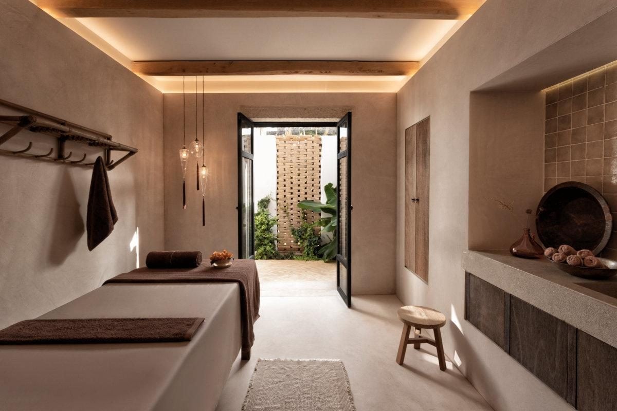 The Rooster, Europe — Spa treatment room with warm earthy tones, indirect ceiling light, massage bed, terrazzo sink, and open doors to brick courtyard garden.