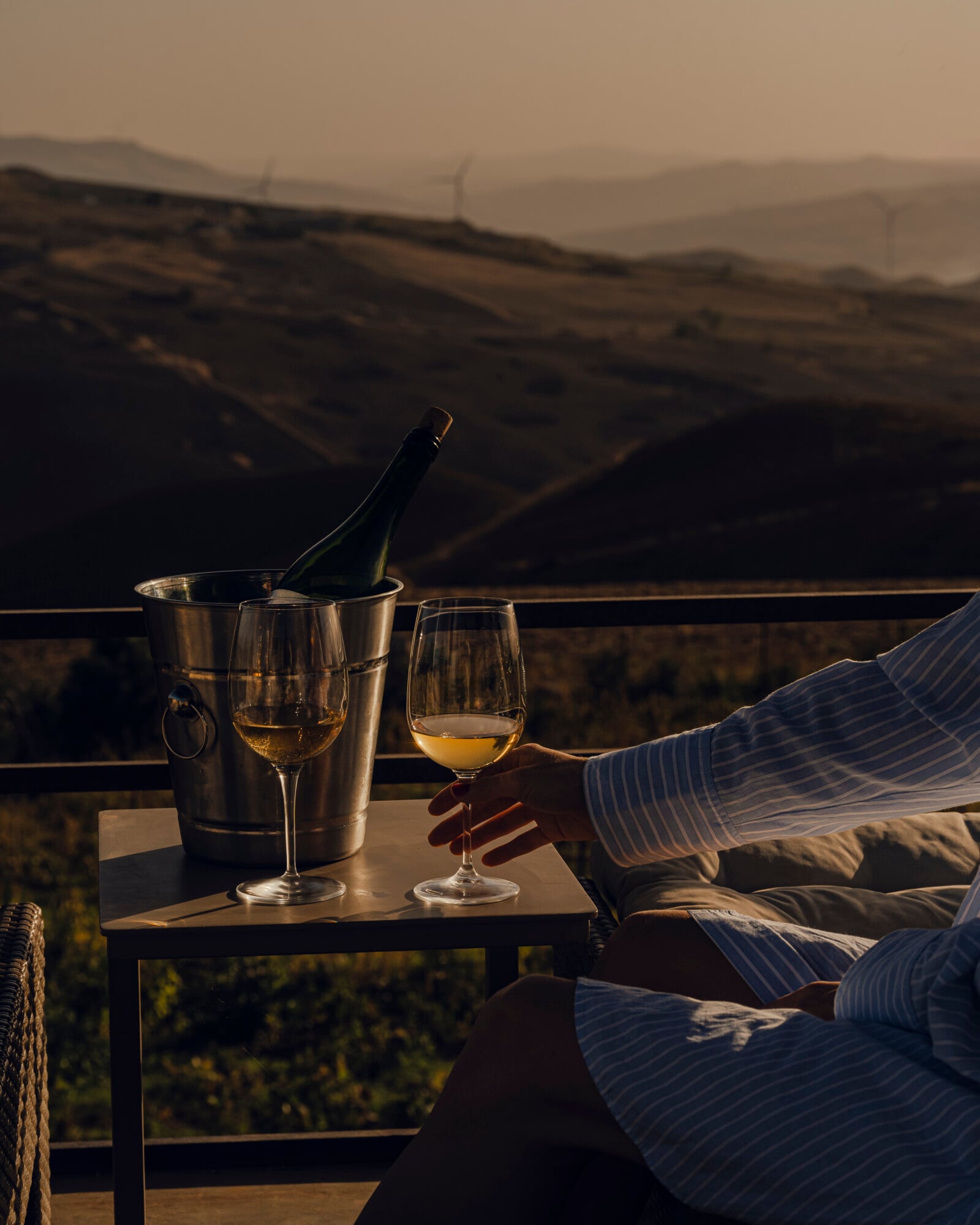 Sicilian Wine Tasting, Italy — Person holding white wine glass on terrace overlooking rolling vineyards at golden hour, metal ice bucket with bottle beside.