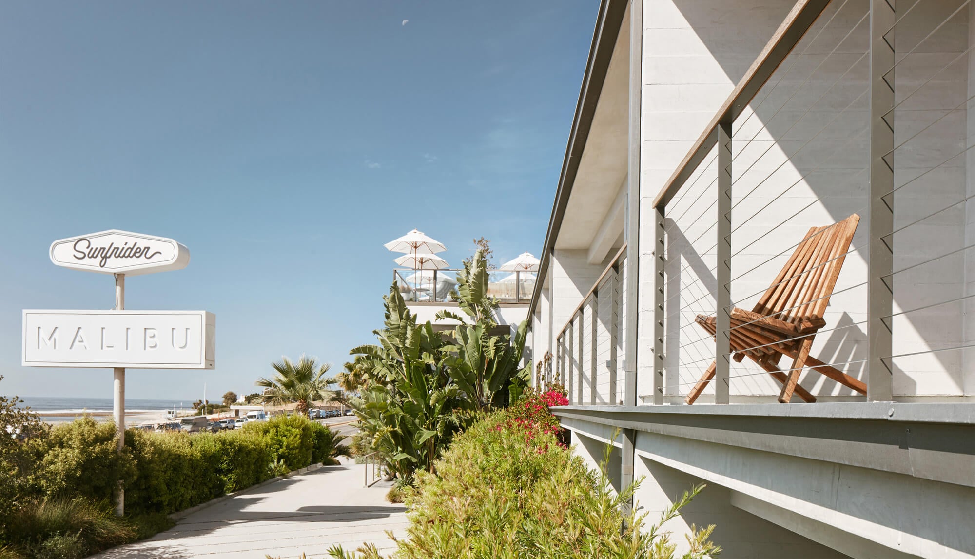 The Surfrider — Surfrider Malibu, California, USA — Modernist beachfront pavilion with white timber louvers, wooden deck chair, and ocean views under clear sky.