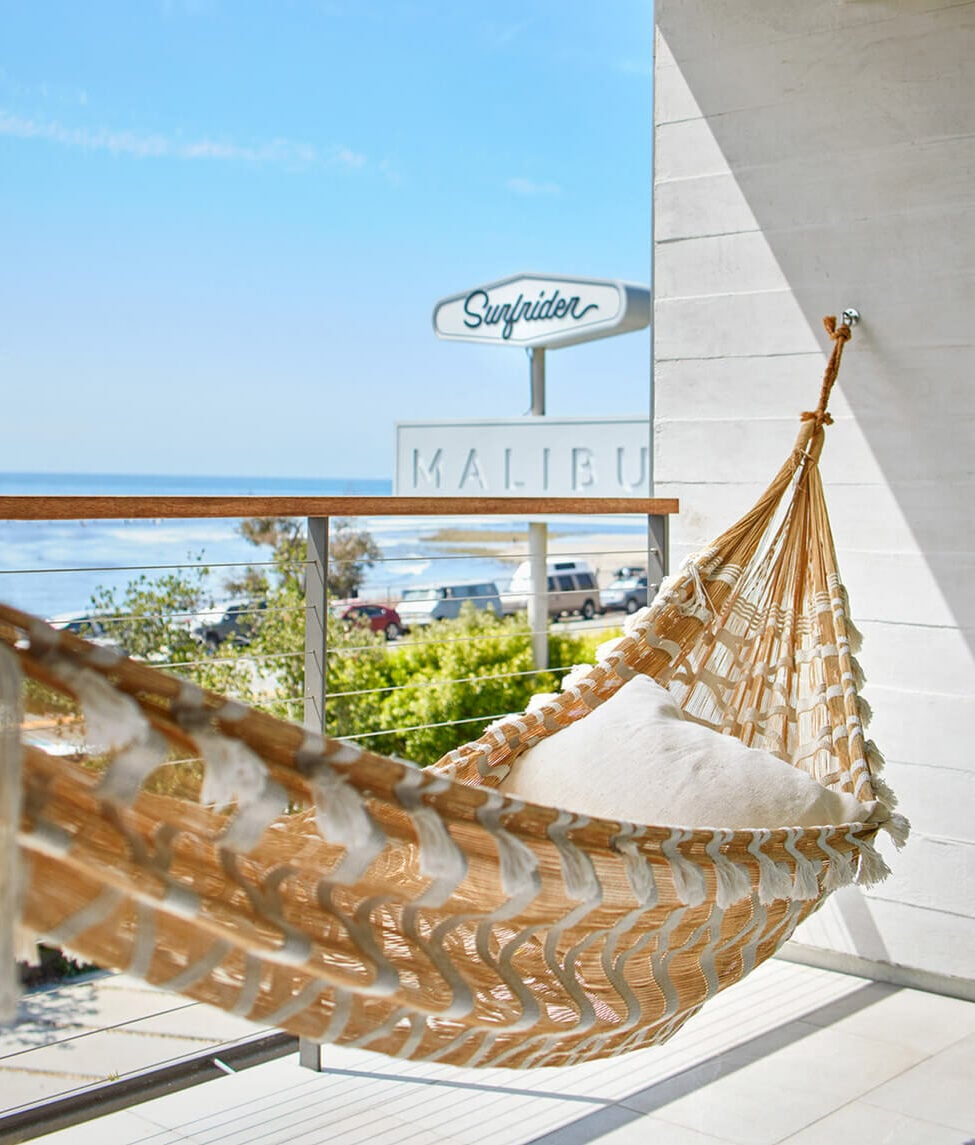 The Surfrider, Malibu, USA — Woven hammock with geometric pattern on coastal terrace overlooking ocean, mid-century modern signage and white architecture.