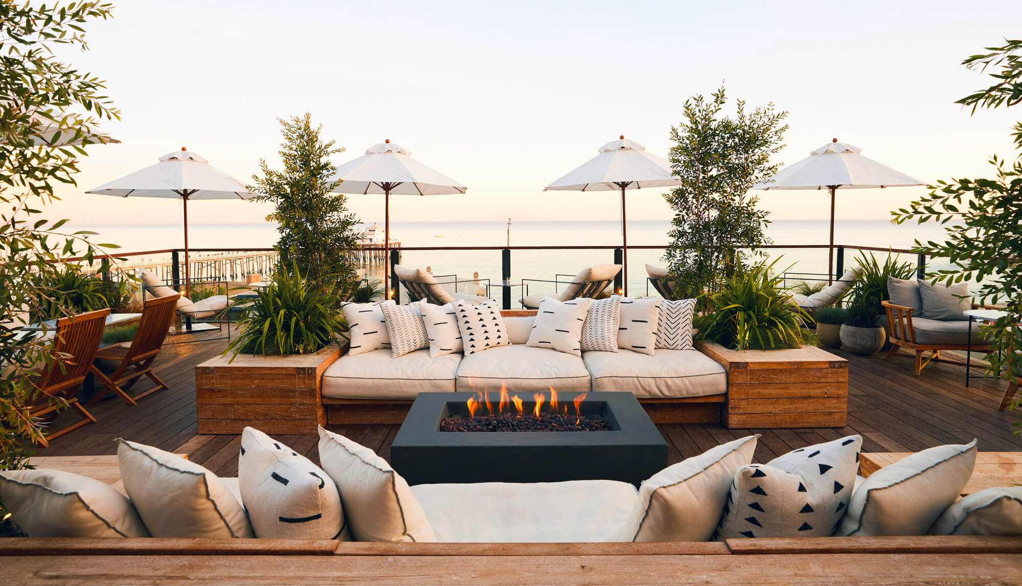 The Surfrider, North America — Oceanfront terrace at sunset with fire pit, teak seating, linen cushions, white umbrellas, and coastal views.