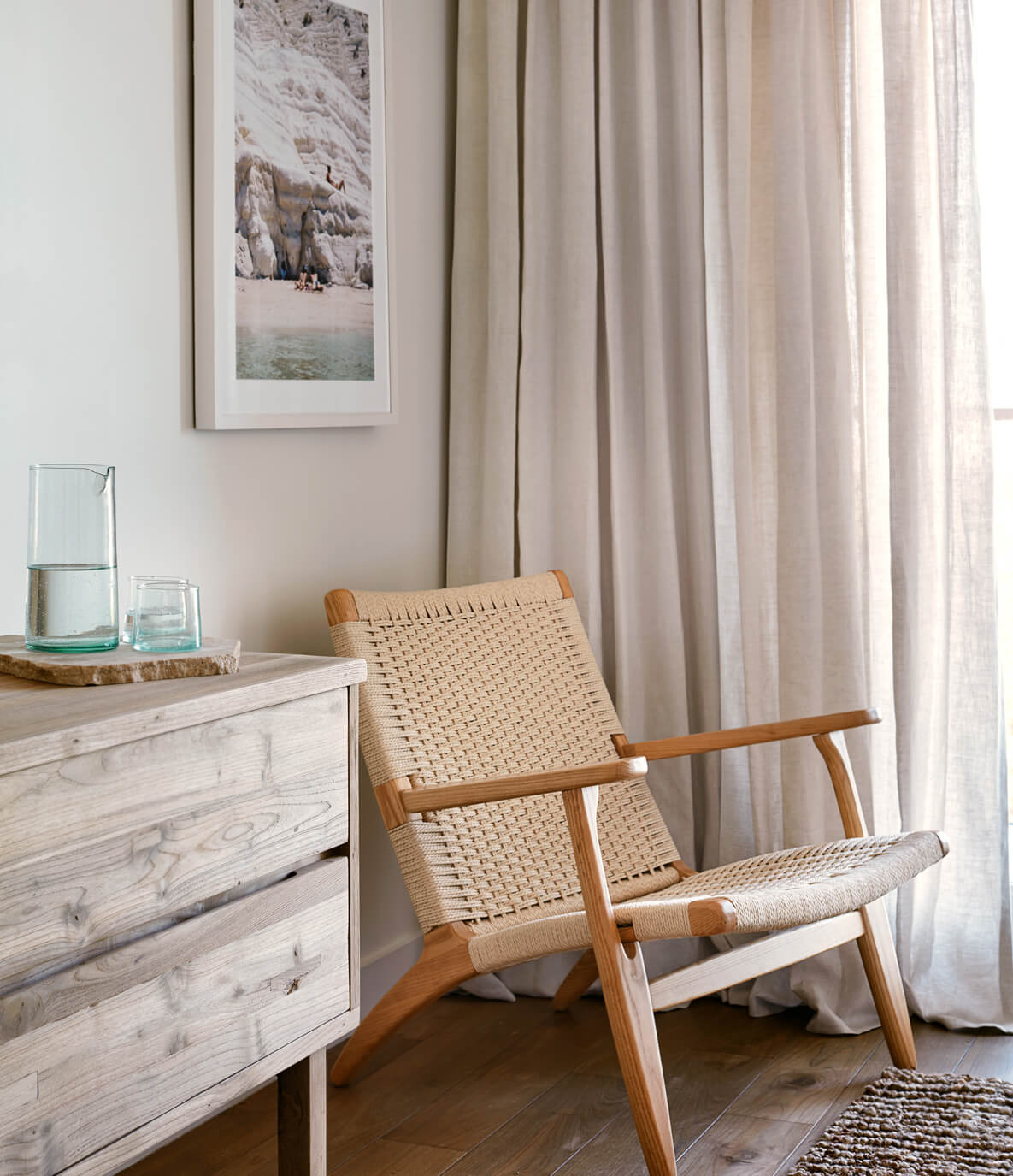 The Surfrider, North America — Coastal modernist bedroom with woven lounge chair, light oak dresser, linen curtains, and framed beach landscape artwork.