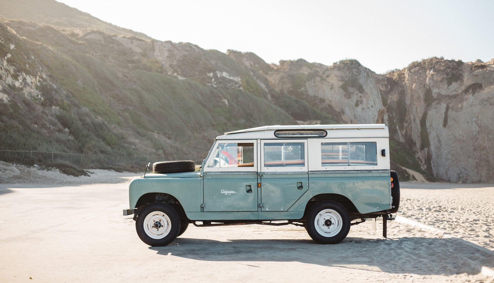 The Surfrider — Vintage Land Rover Series III parked on sandy beach with white chalk cliffs rising behind, golden hour light, coastal landscape.