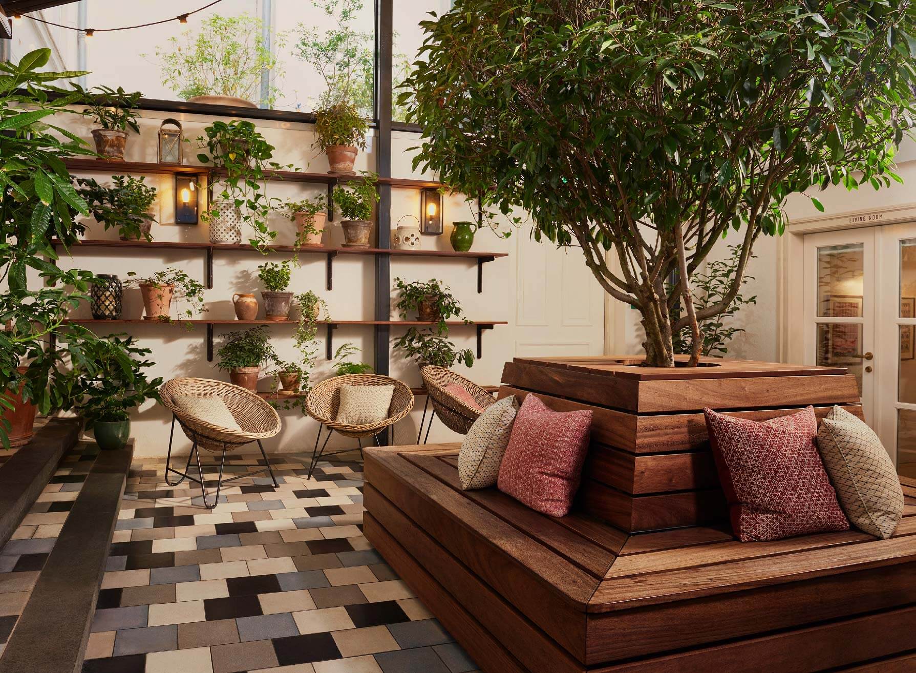 The Hoxton Poblenou, Barcelona, Spain — Plant-filled conservatory with wooden shelving, woven seating, and built-in sofa beside large tree planter, geometric tile flooring, warm ambient lighting.
