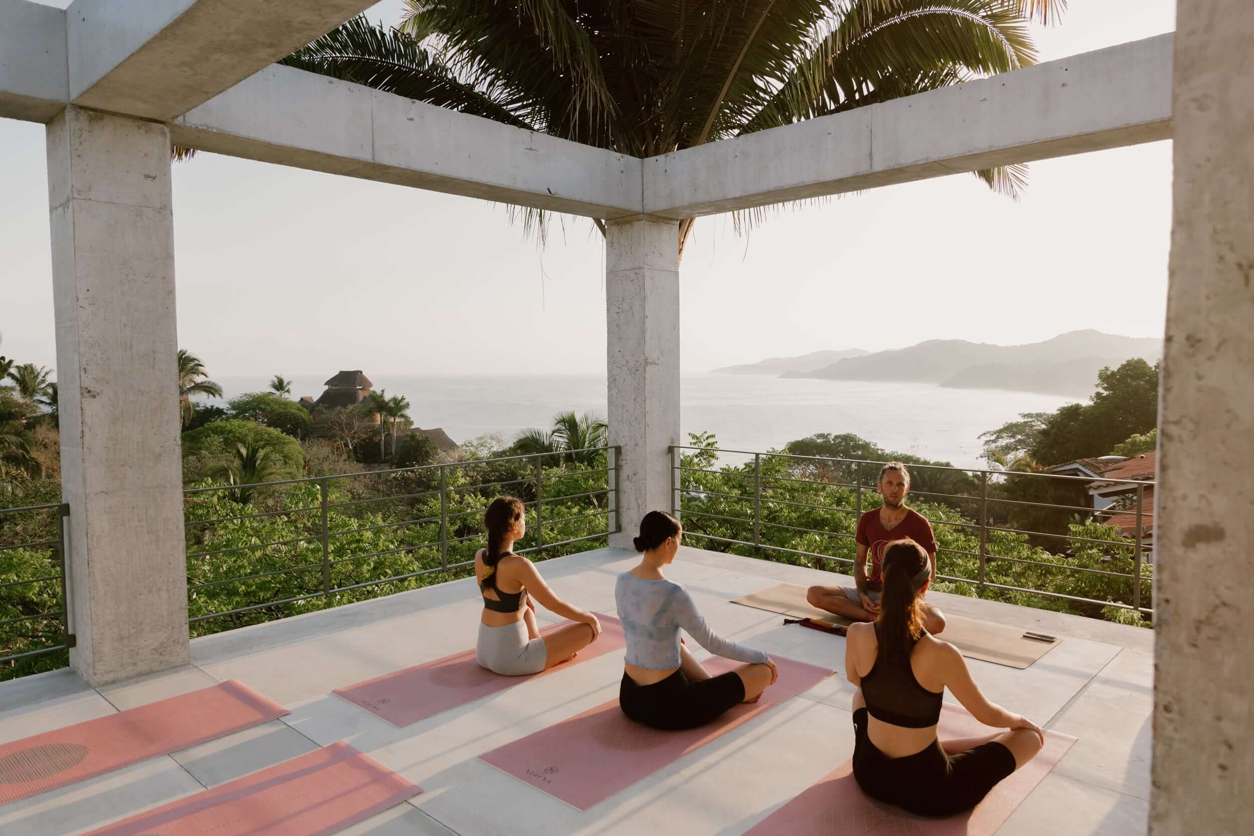 Rooftop Yoga, Mexico — Instructor leading three students in seated poses on coral mats beneath concrete pergola with palm fronds, ocean and mountains visible beyond metal railings at sunset.
