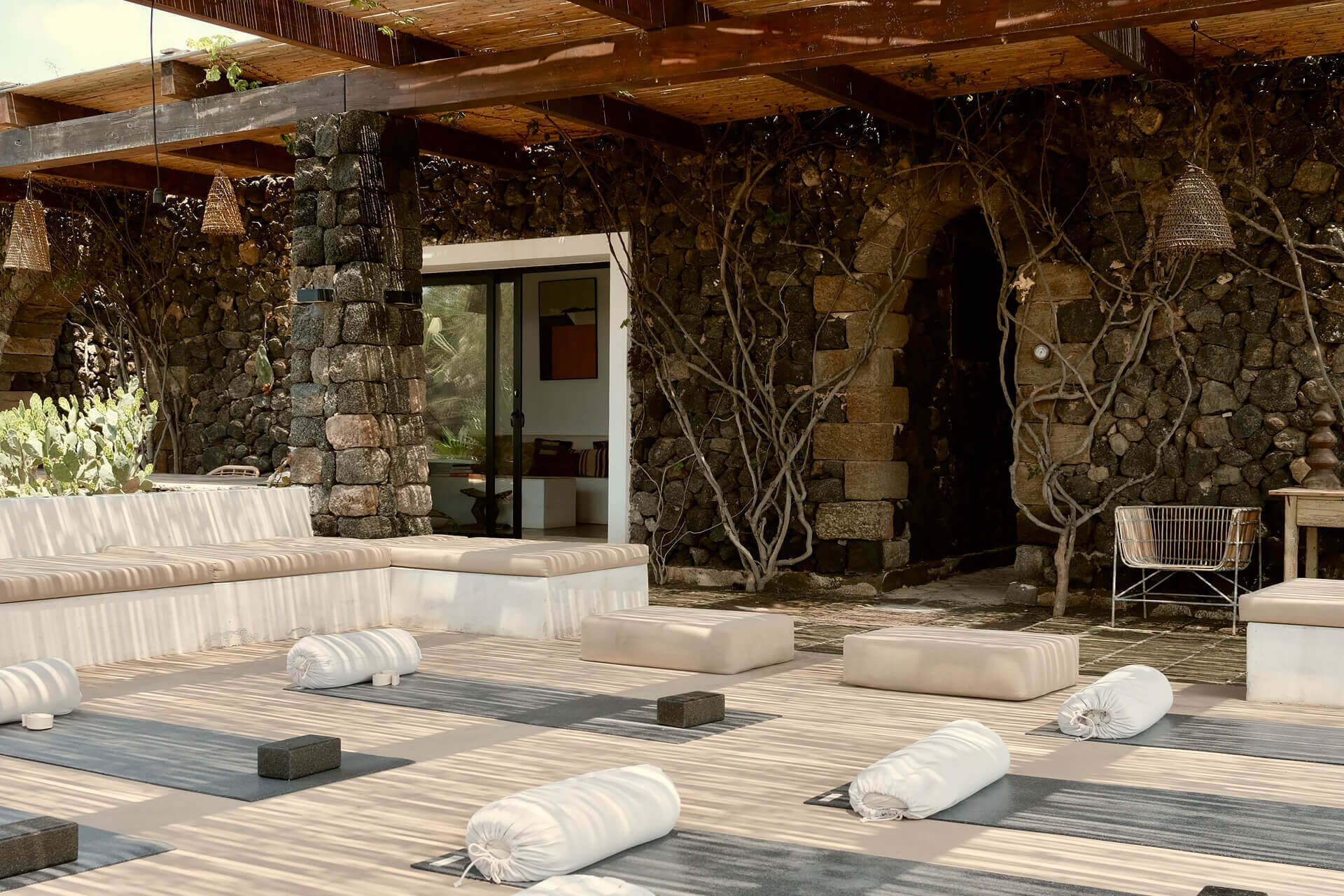 Parco dei Sesi Retreat, Europe — Open-air yoga studio with stone walls, wooden beams, rolled white cushions on natural wood flooring, grapevine-draped architecture, minimalist hospitality design.