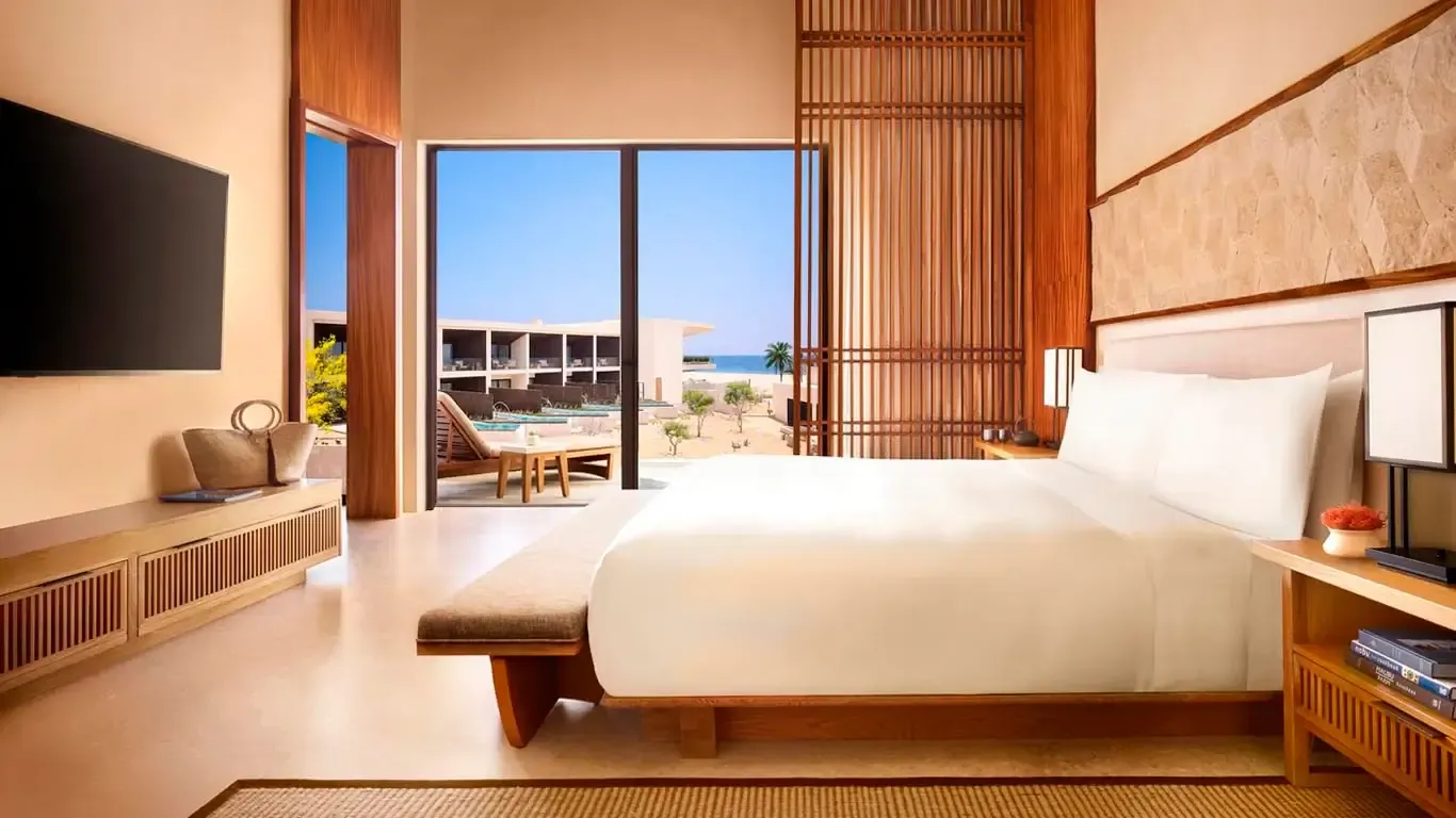 Nobu Los Cabos, Mexico — Modern beachfront bedroom with warm wood paneling, minimalist white bedding, floor-to-ceiling ocean views, and contemporary coastal design aesthetic.