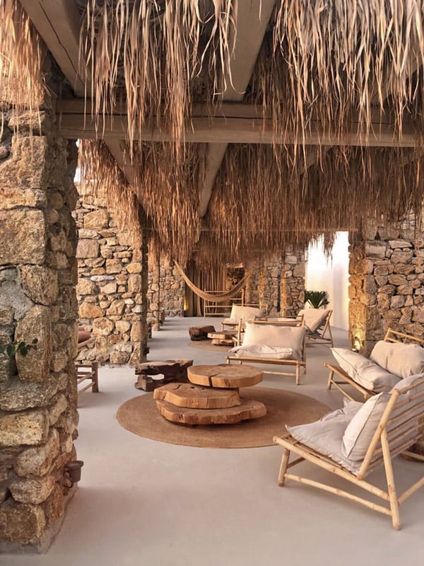 The Wild, Europe — Thatched pergola pavilion with stone columns, wooden loungers, woven palm ceiling, central fire pit, warm natural light.