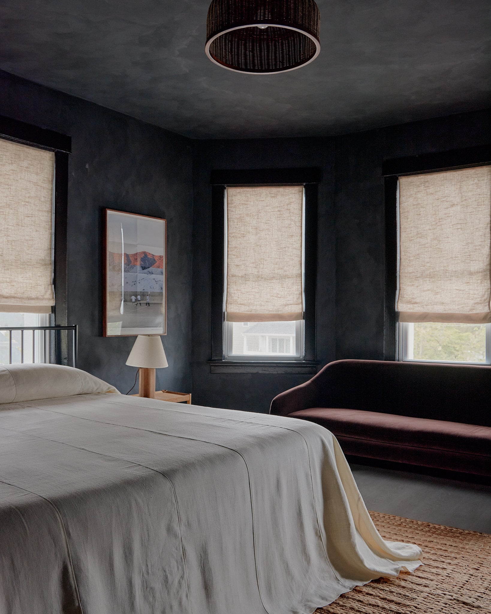 The James Bradley, North America — Dark concrete bedroom with linen roman shades, wooden nightstand lamp, burgundy velvet sofa, woven area rug, and mountain landscape artwork.