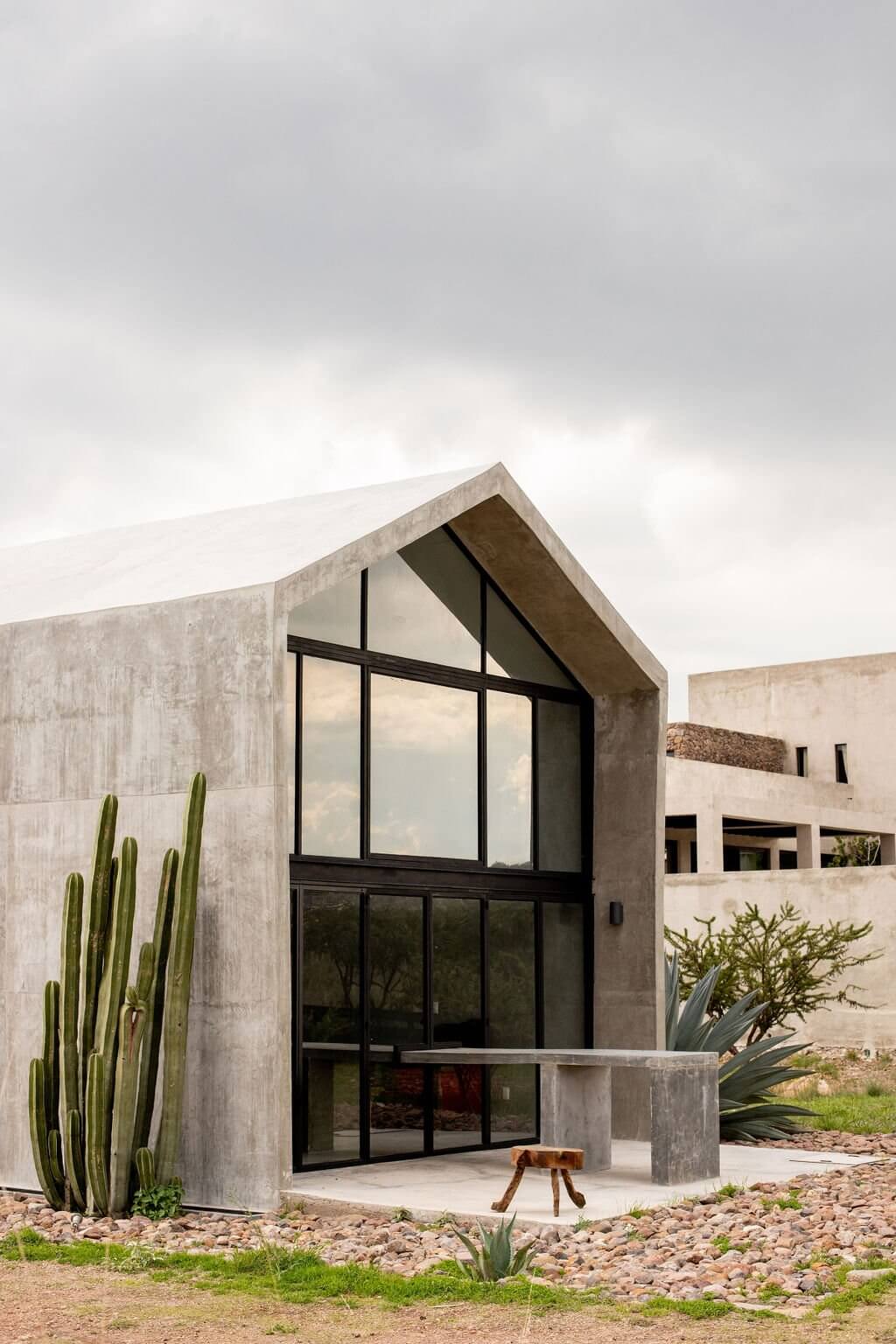 Villa ITO, Mexico — Minimalist concrete pavilion with black-framed glazing, pitched roof, desert landscaping, and stone terrace under overcast sky.