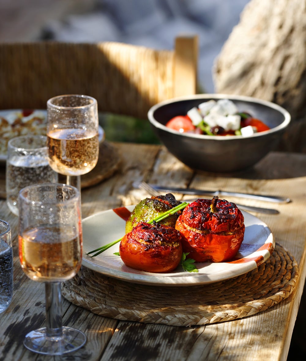 Taverna, Europe — Charred red peppers with green onion on white plate, sparkling wine glasses, salad bowl, rustic wooden table in Mediterranean sunlight.