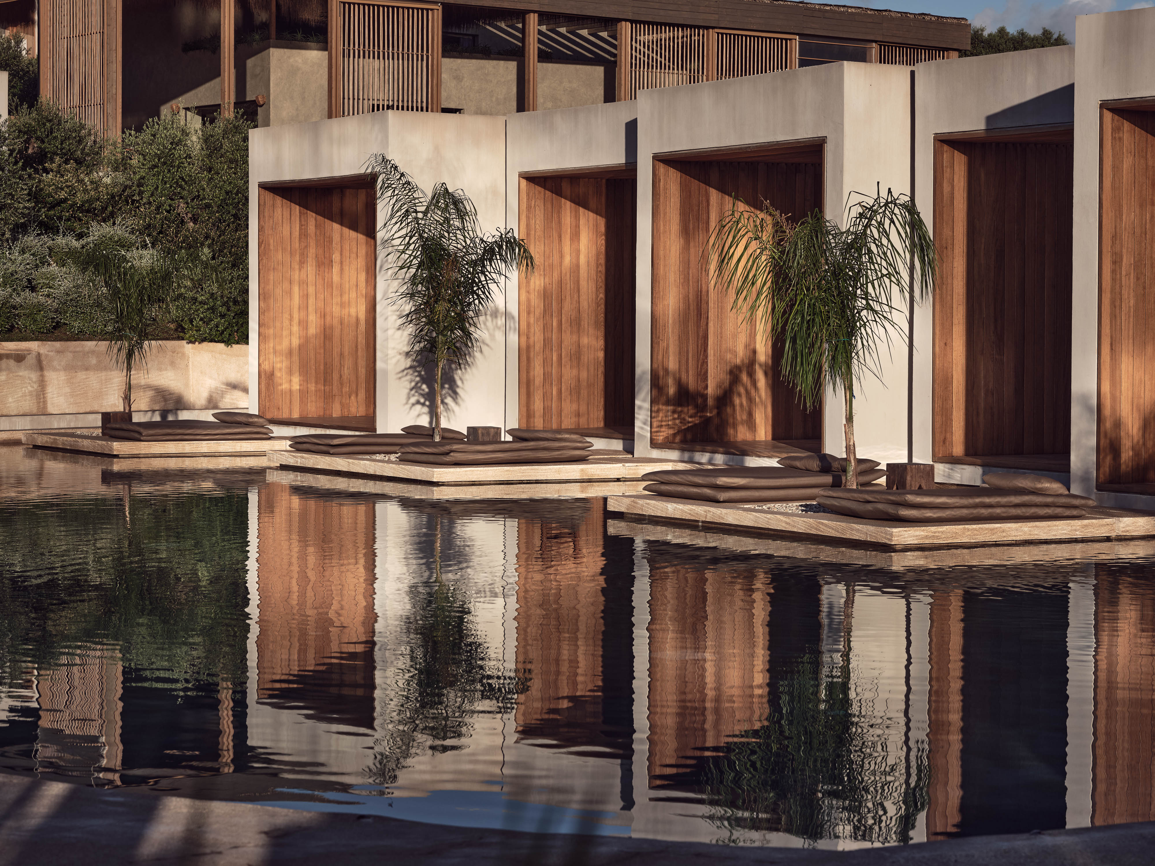 Olea All Suite Hotel, Europe — Minimalist poolside pavilions with timber doors, cream walls, and cascading palms reflected in still water at golden hour.
