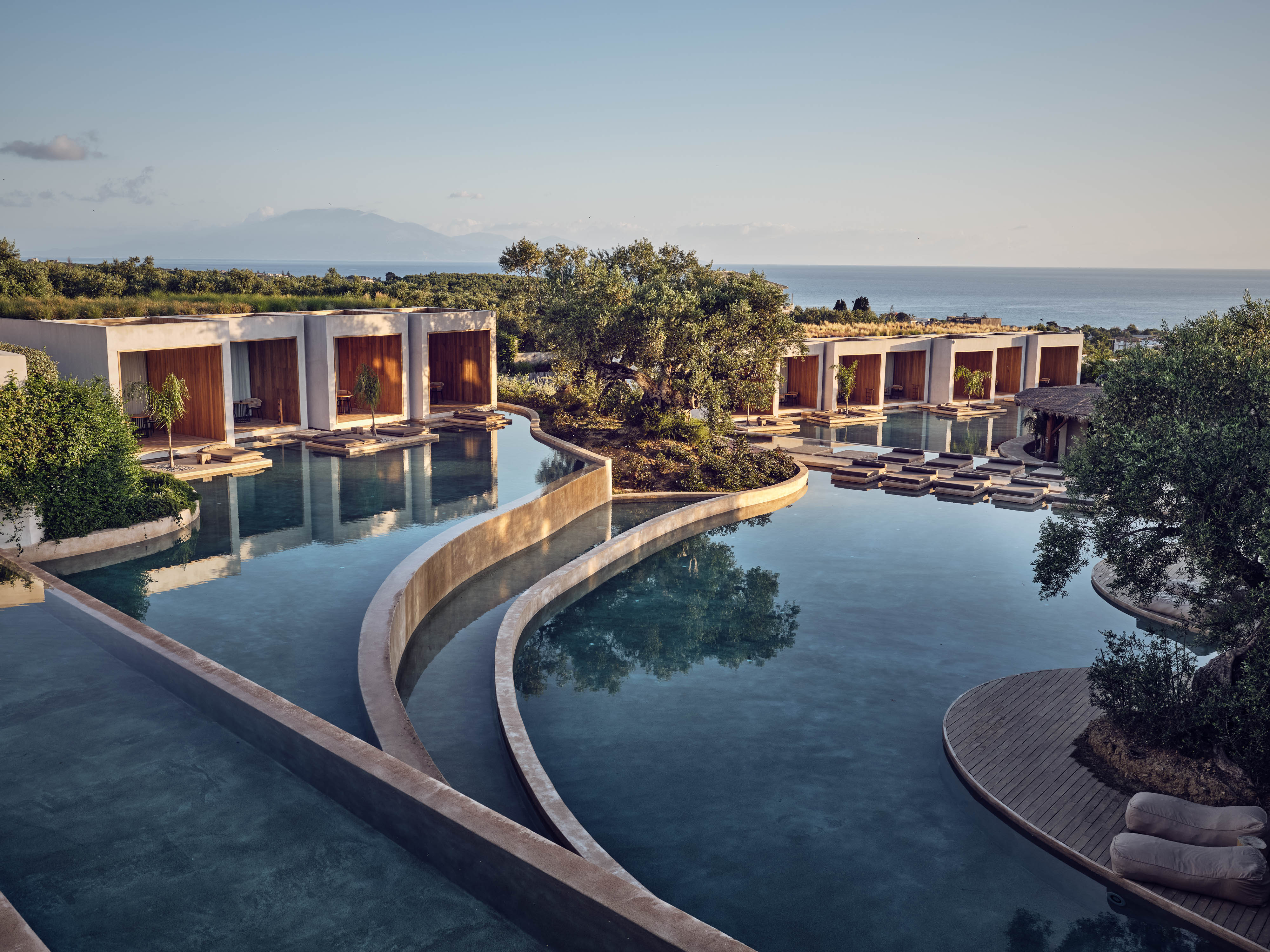Olea All Suite Hotel, Europe — Curved concrete poolside pathways connect modernist pavilions with terracotta shutters, overlooking sea and mountain landscape at dusk.