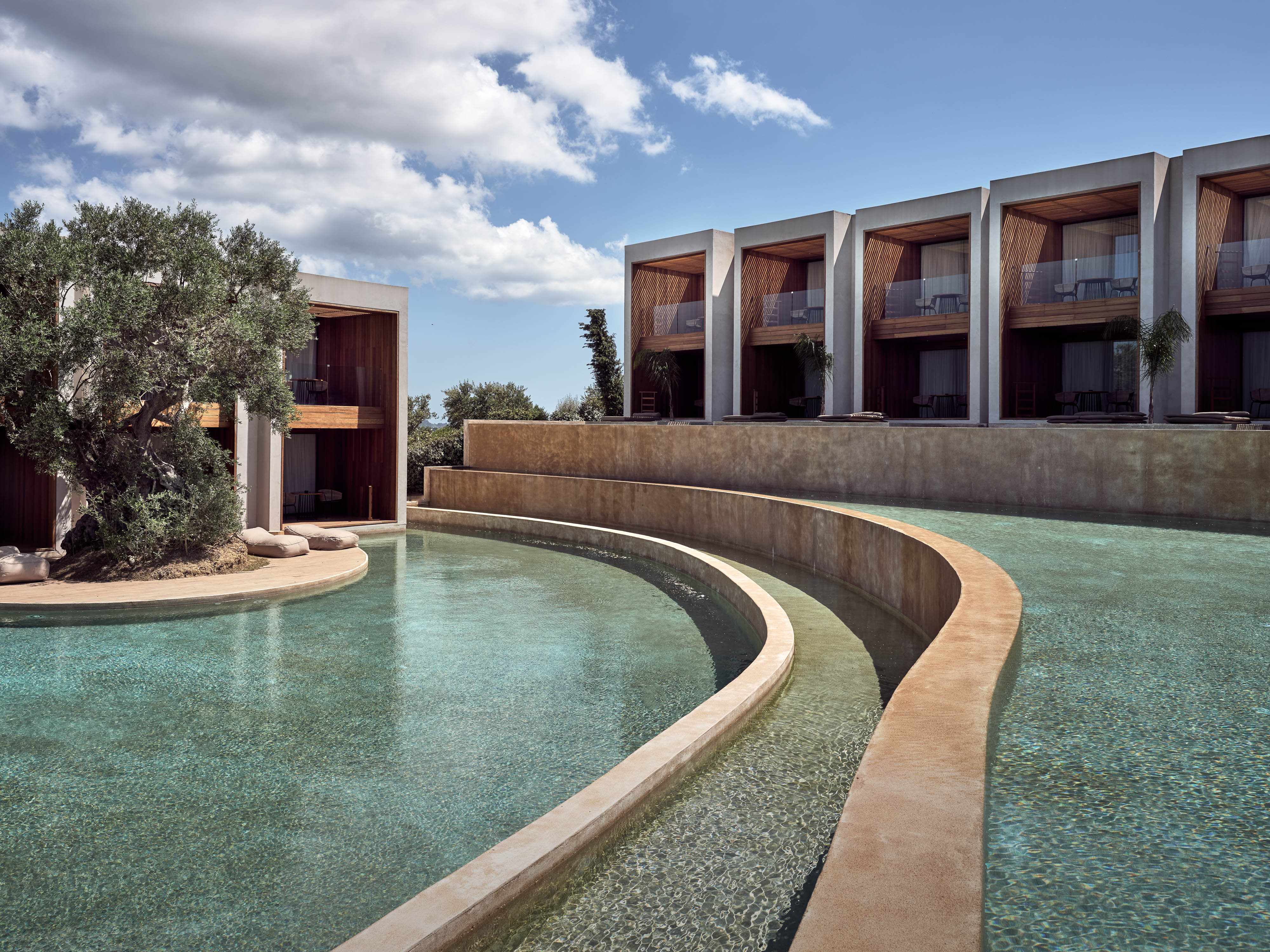 Olea All Suite Hotel, Europe — Curved waterway reflects turquoise light beside modernist white pavilions with wooden screens and olive tree courtyard.