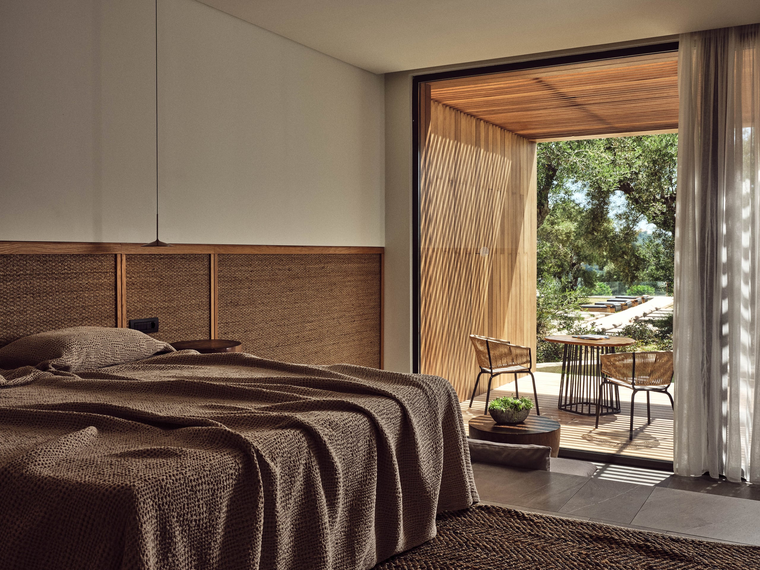 Olea All Suite Hotel, Europe — Minimalist bedroom with woven headboard, natural linen bedding, wooden ceiling, and terrace overlooking landscaped gardens and pool.