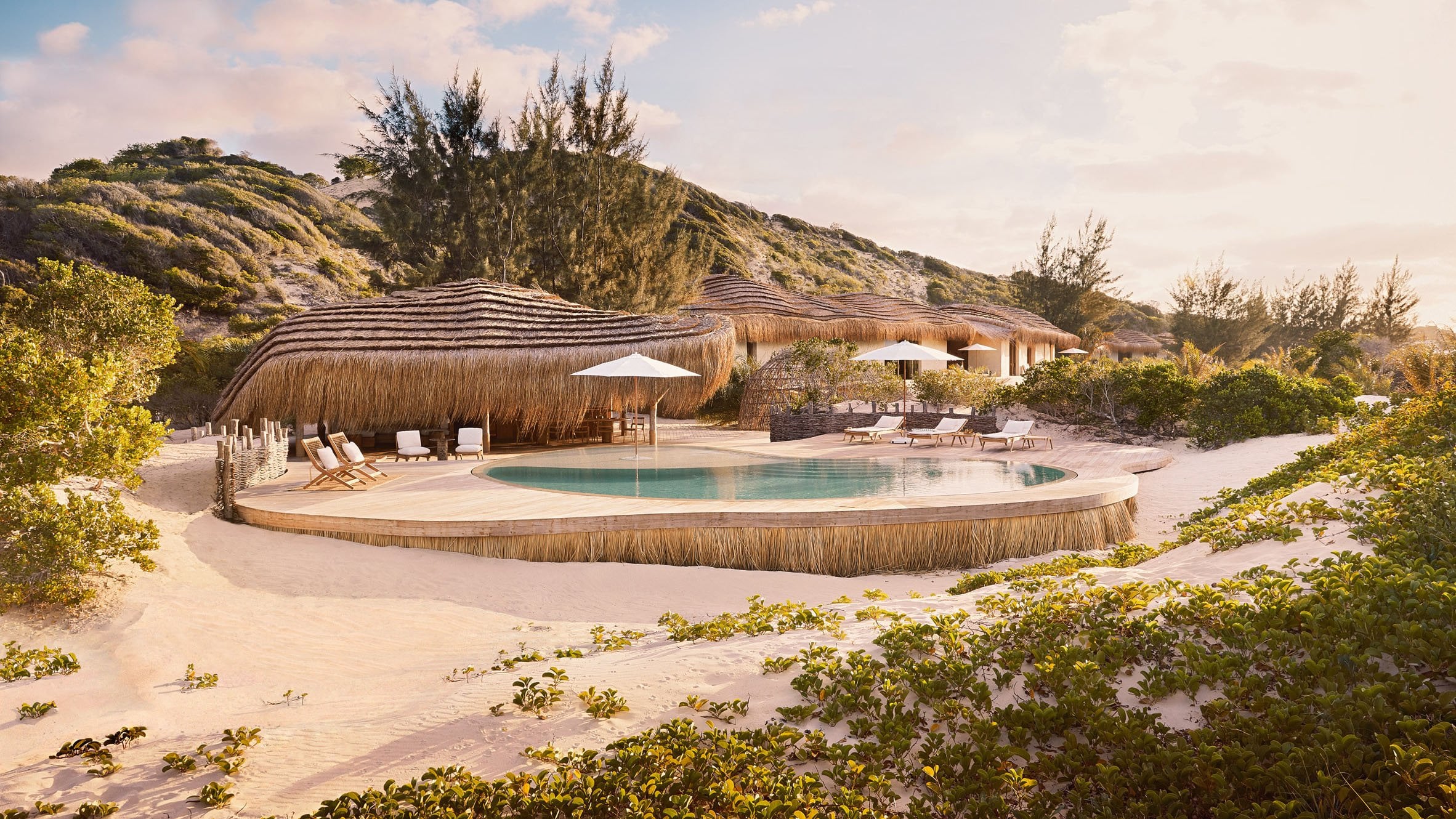 Kisawa Sanctuary, Africa — Thatched-roof pavilions with curved architecture overlook turquoise pool surrounded by sand, native vegetation, and rocky hills at dusk.