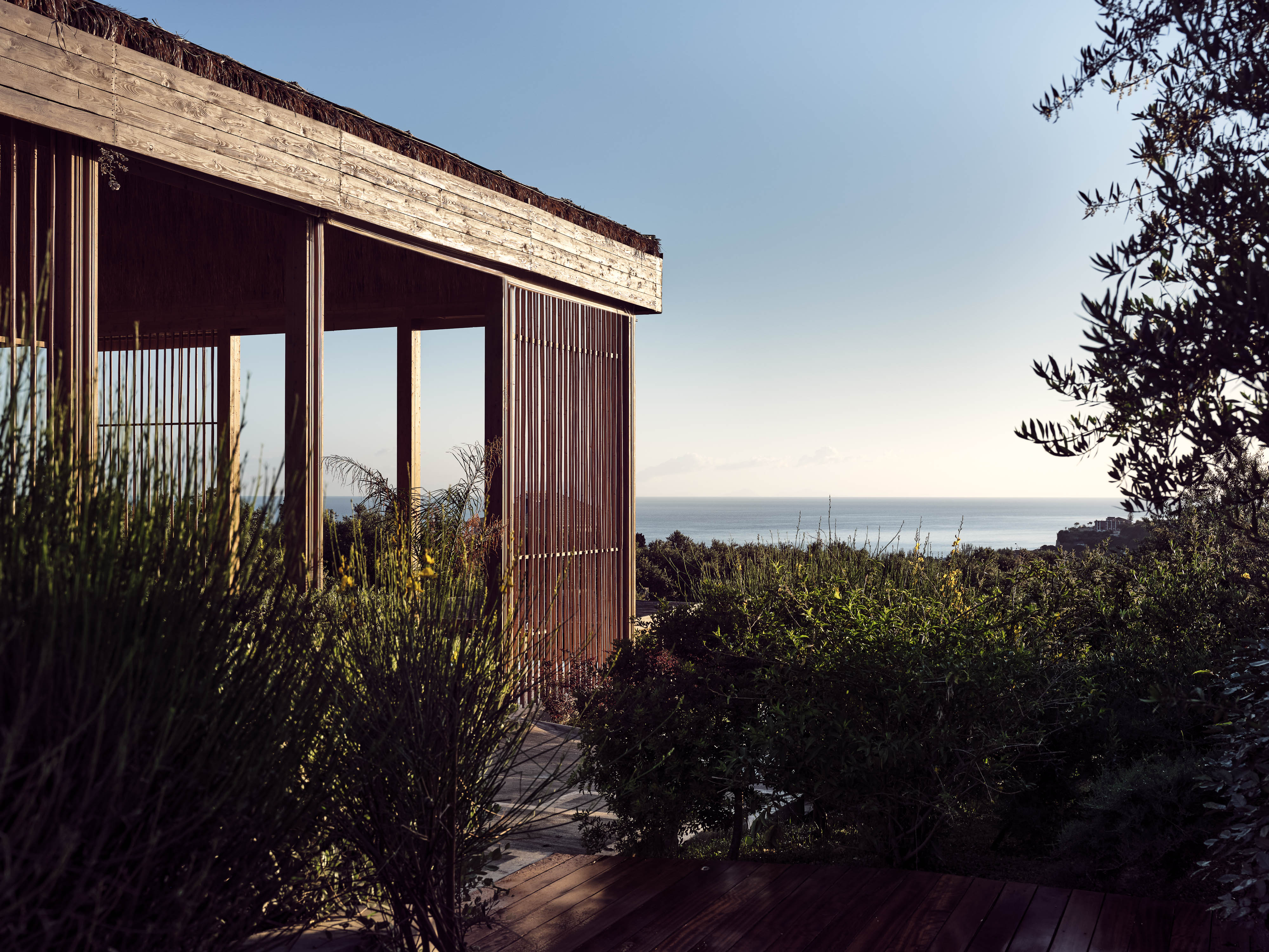 Olea All Suite Hotel, Europe — Modern wooden pavilion with vertical slat screens framing Mediterranean seascape through open glass doors, surrounded by native vegetation and warm stone terrace.