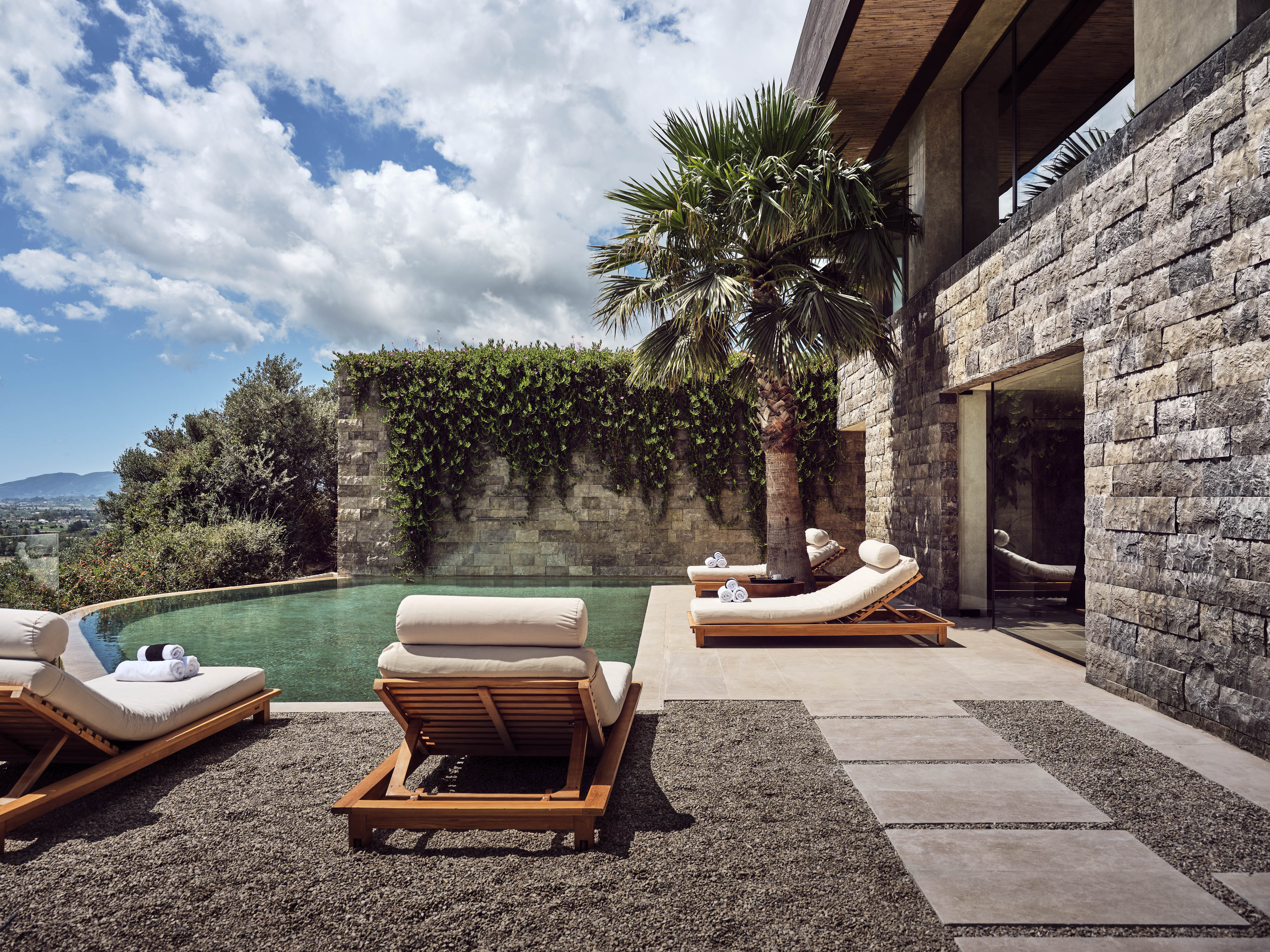 Olea All Suite Hotel, Europe — Limestone terrace with heated pool, teak loungers, ivy-covered stone wall, palm tree, and mountain views under blue sky.