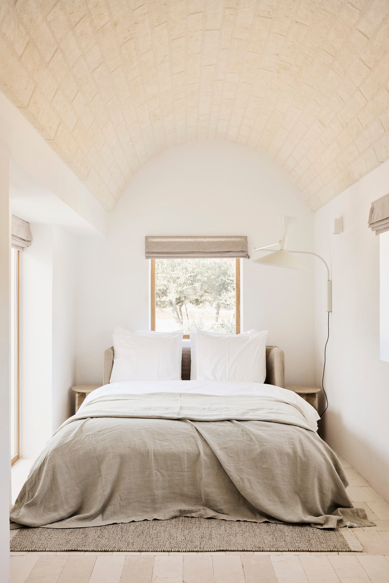 Villa Sessana, Italy — Minimalist bedroom with vaulted brick ceiling, neutral linens, wooden-framed window overlooking trees, natural light, concrete floor with woven rug.
