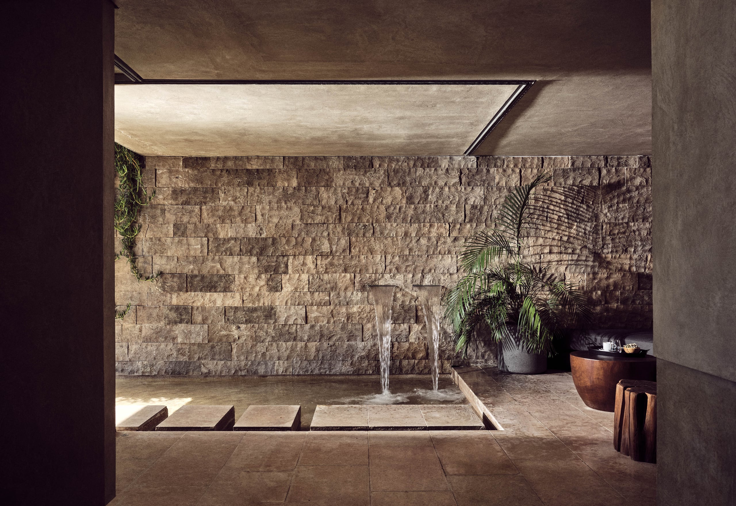 Olea All Suite Hotel, Europe — Minimalist courtyard with cascading water feature, textured stone walls, potted palms, and skylight flooding concrete surfaces with natural light.