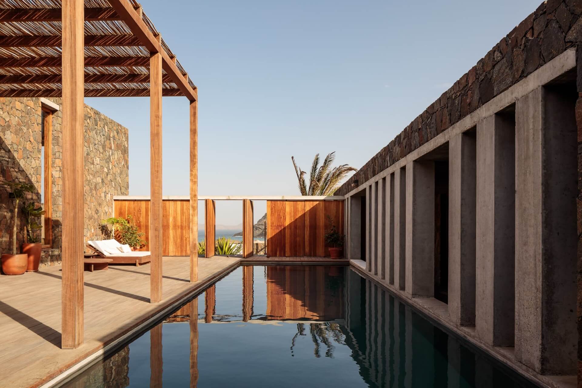 Barefoot Luxury, Africa — Minimalist wooden pergola and reflecting pool between stone and concrete structures overlooking coastal landscape at sunset.