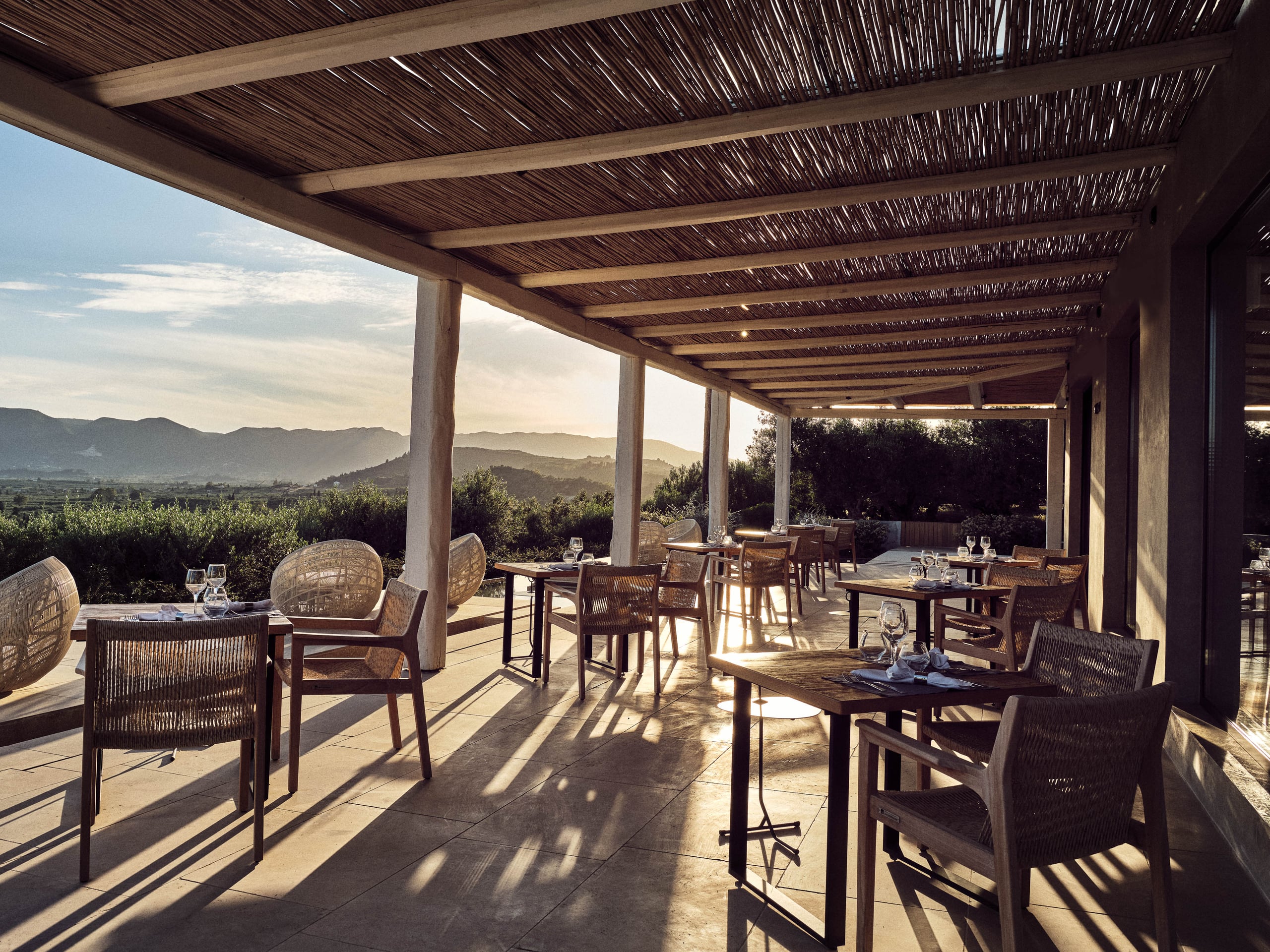 Olea All Suite Hotel, Europe — Pergola-shaded terrace with woven roof panels, wooden dining furniture, and mountain valley views at golden hour.