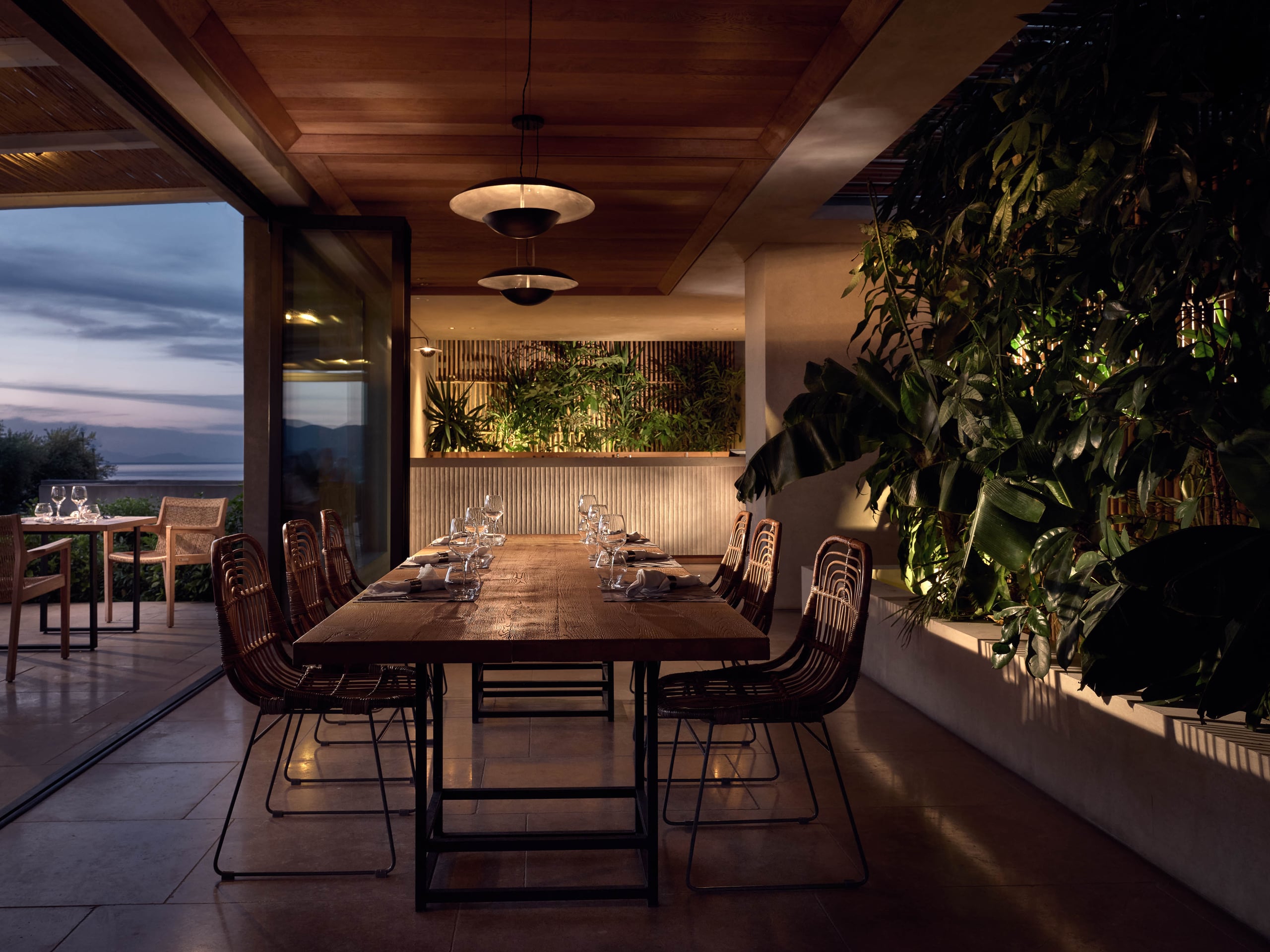 Olea All Suite Hotel, Europe — Modern dining pavilion at dusk with timber ceiling, rattan chairs, living plant wall, and panoramic water views through glass doors.