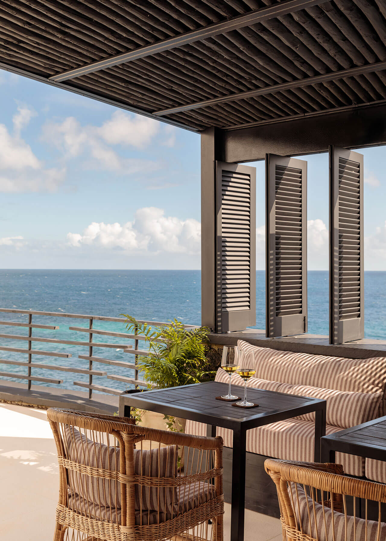 White Hotel, Europe — Oceanfront terrace with louvered shutters, woven seating, black table, and striped cushions beneath slatted pergola overlooking calm blue sea.