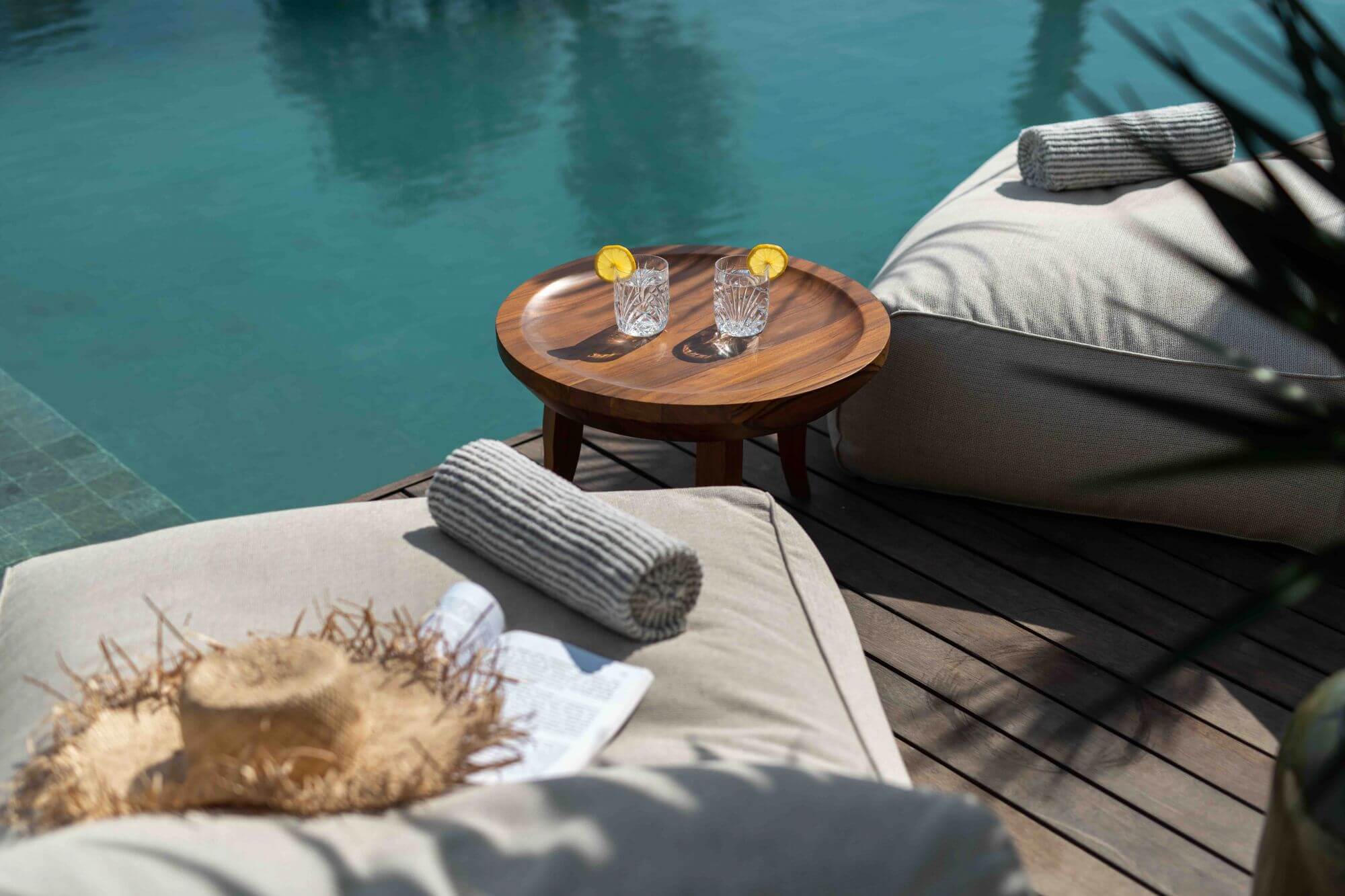 Cap Karoso, Asia — Wooden side table with crystalline glasses beside loungers overlooking turquoise pool, rolled linens and sea urchin sculpture detail.