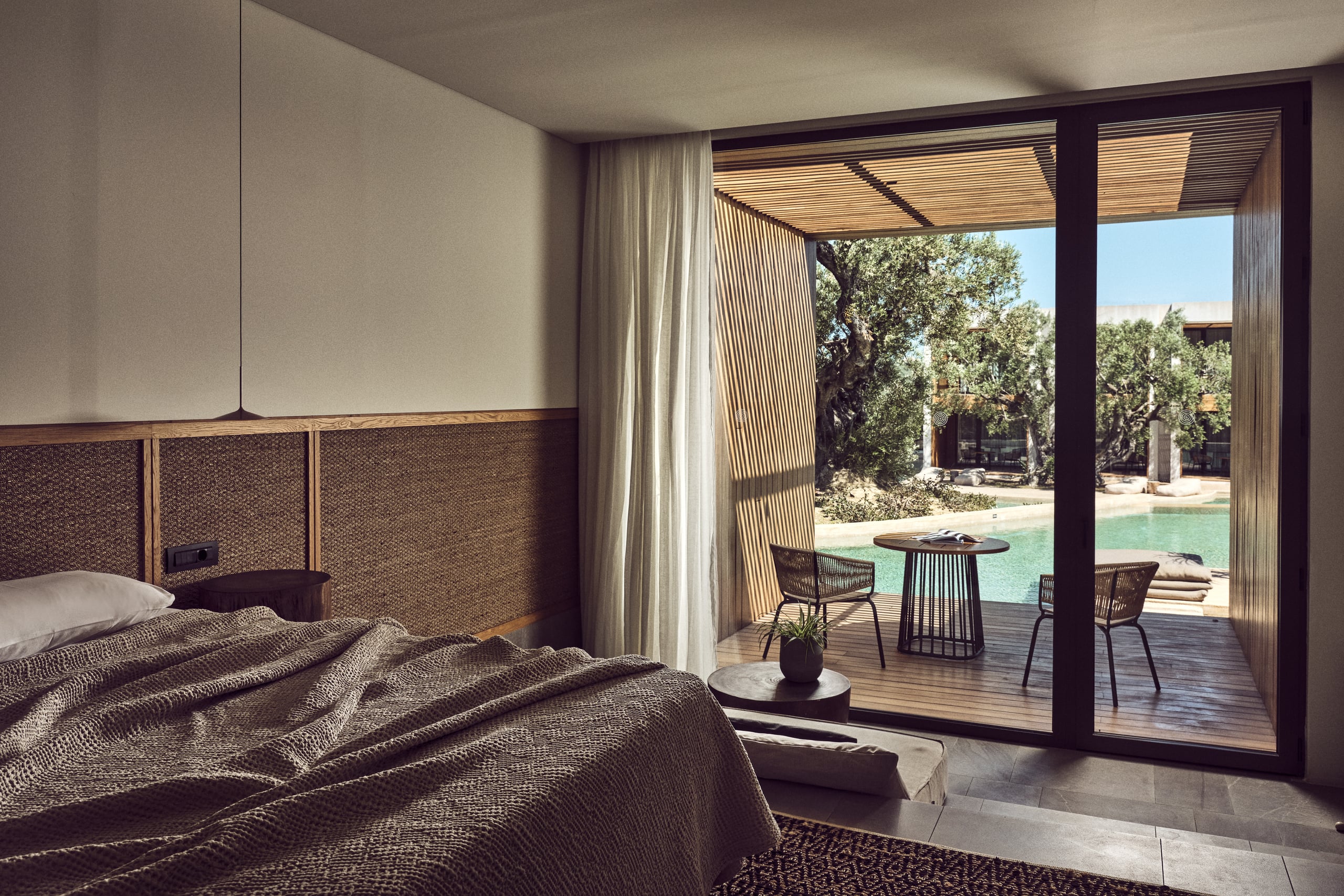 Olea All Suite Hotel, Europe — Bedroom with woven headboard opening to poolside terrace, wooden pergola, and olive trees under clear sky.