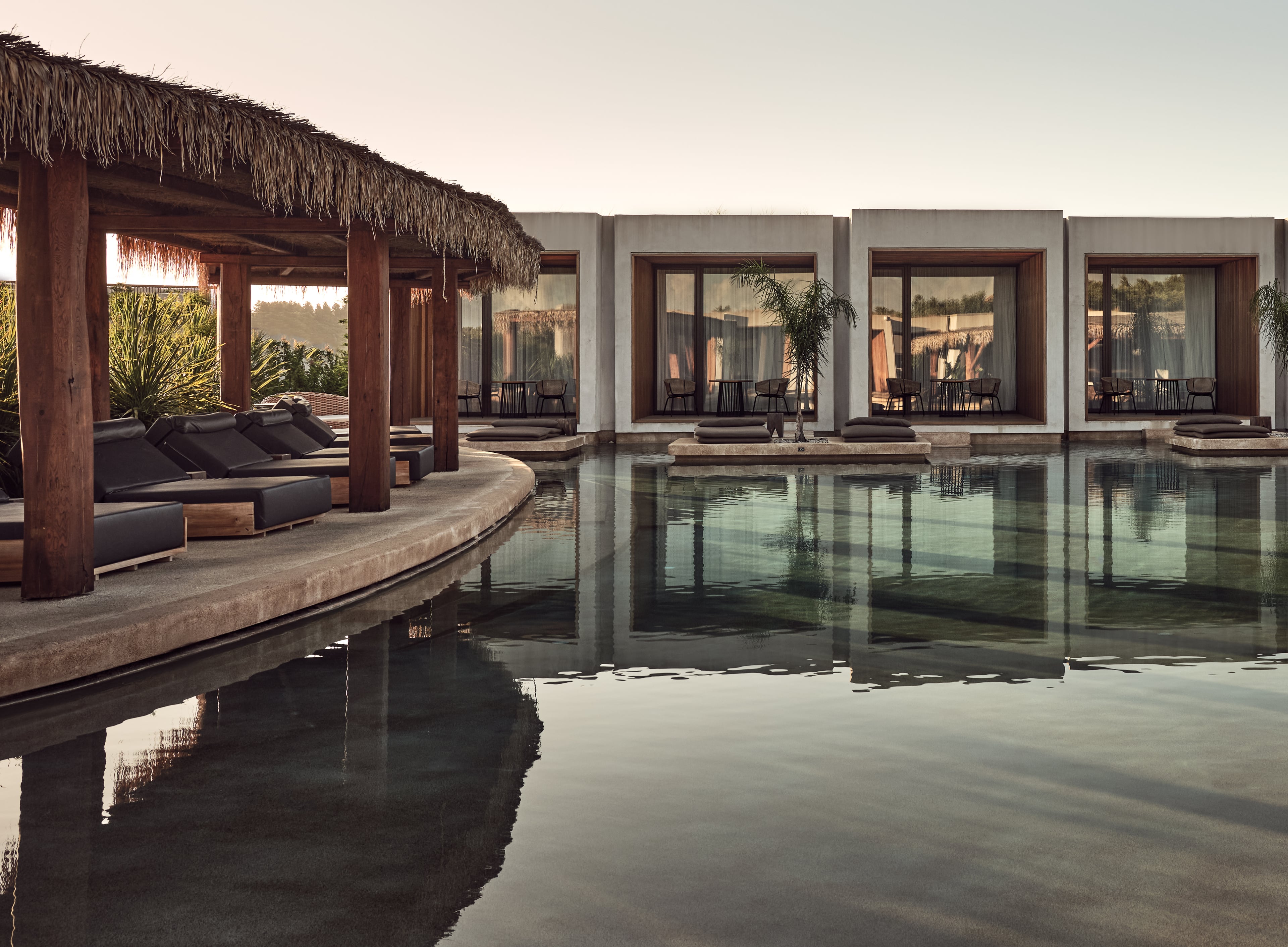Olea All Suite Hotel, Europe — Minimalist poolside terrace with thatched pergola, sunbeds, reflective water feature, and modernist glass-fronted pavilion at golden hour.